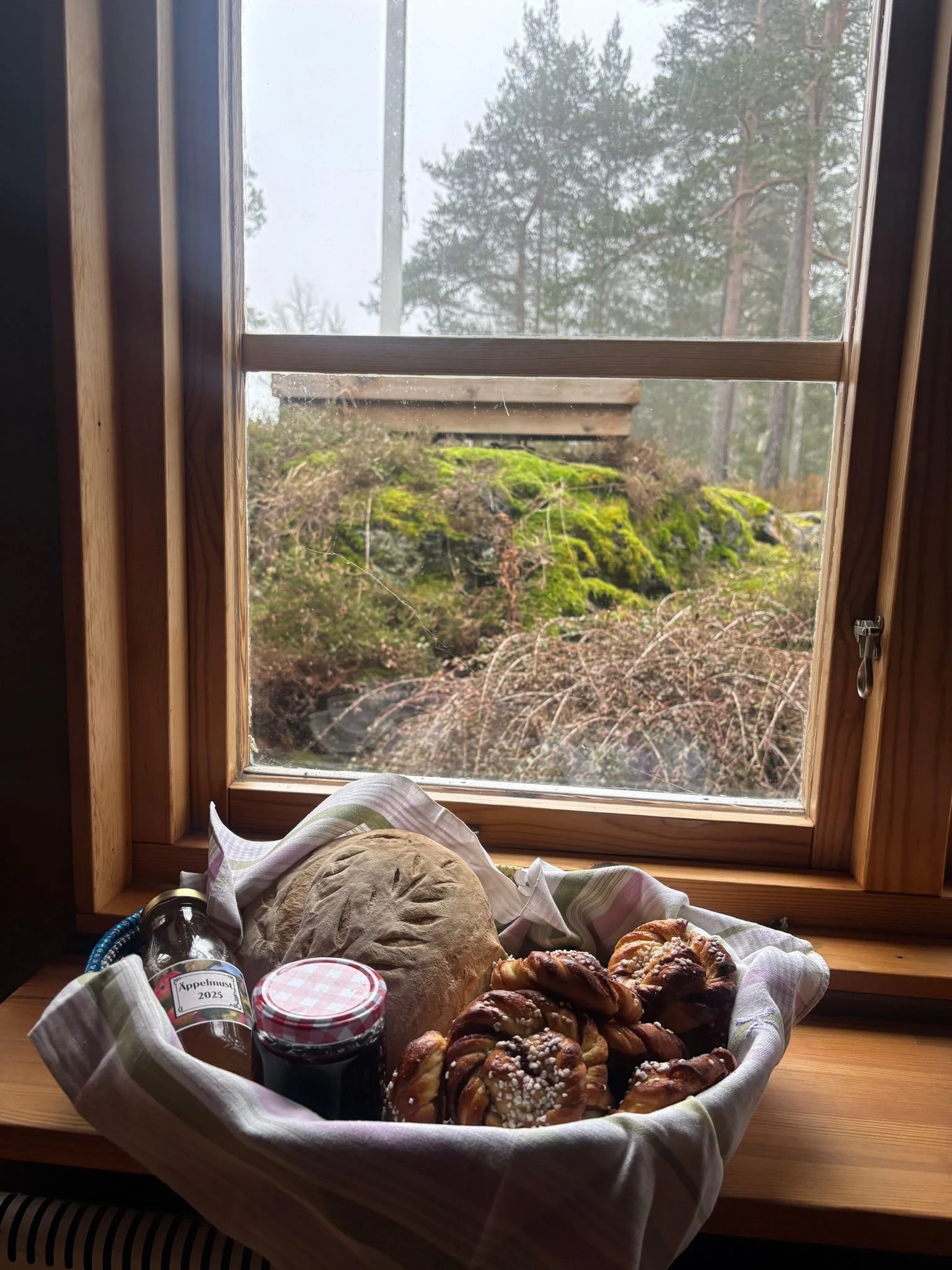 A basket of baked pretzels, a loaf of bread, jars of jam, and a cloth, set on a wooden windowsill with a view of trees outside.