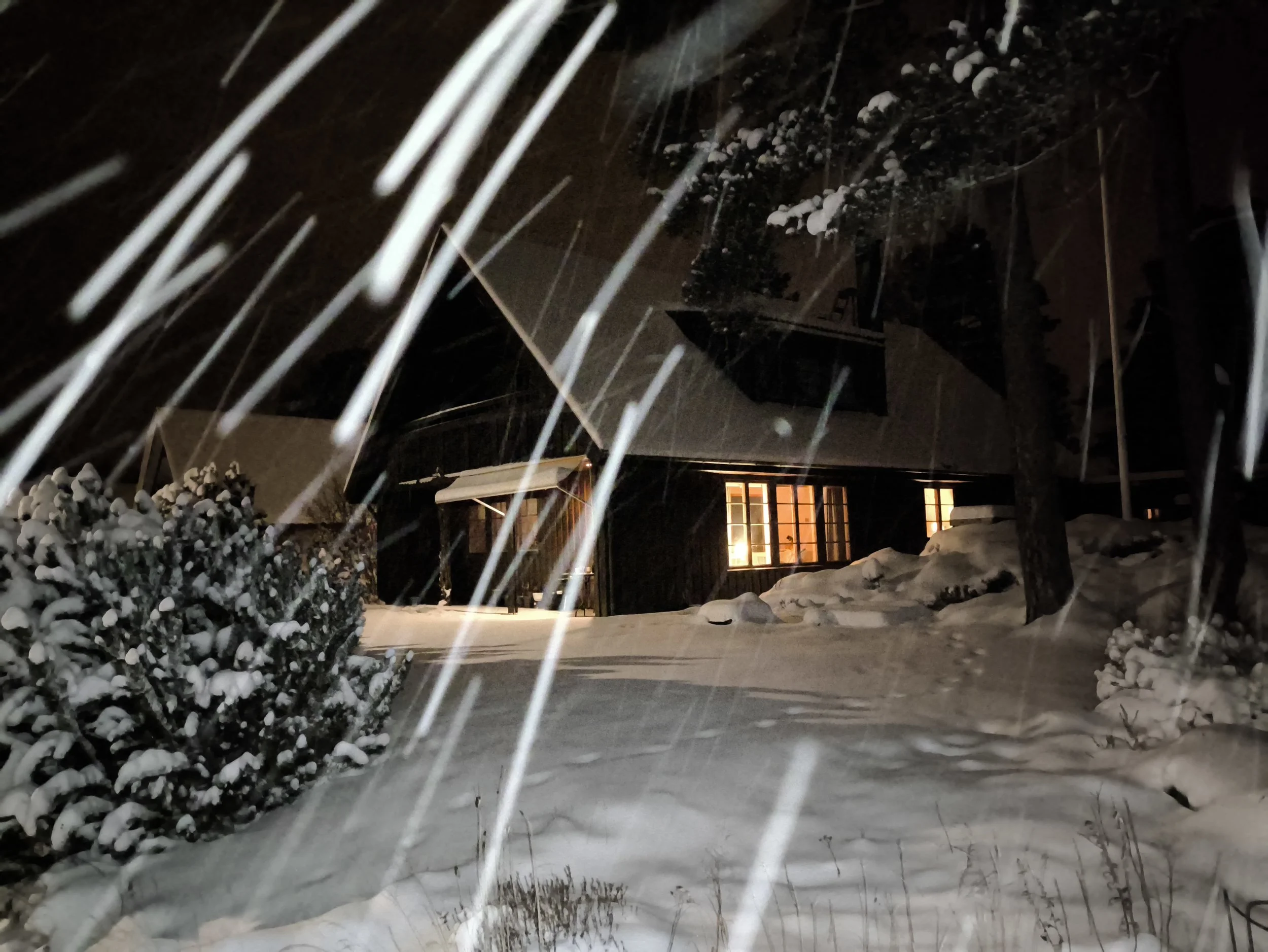 Snow falling overnight on a residential house with illuminated windows, a snow-covered driveway, and snow-laden bushes and trees. West Cottage Sweden, Åsensbruk