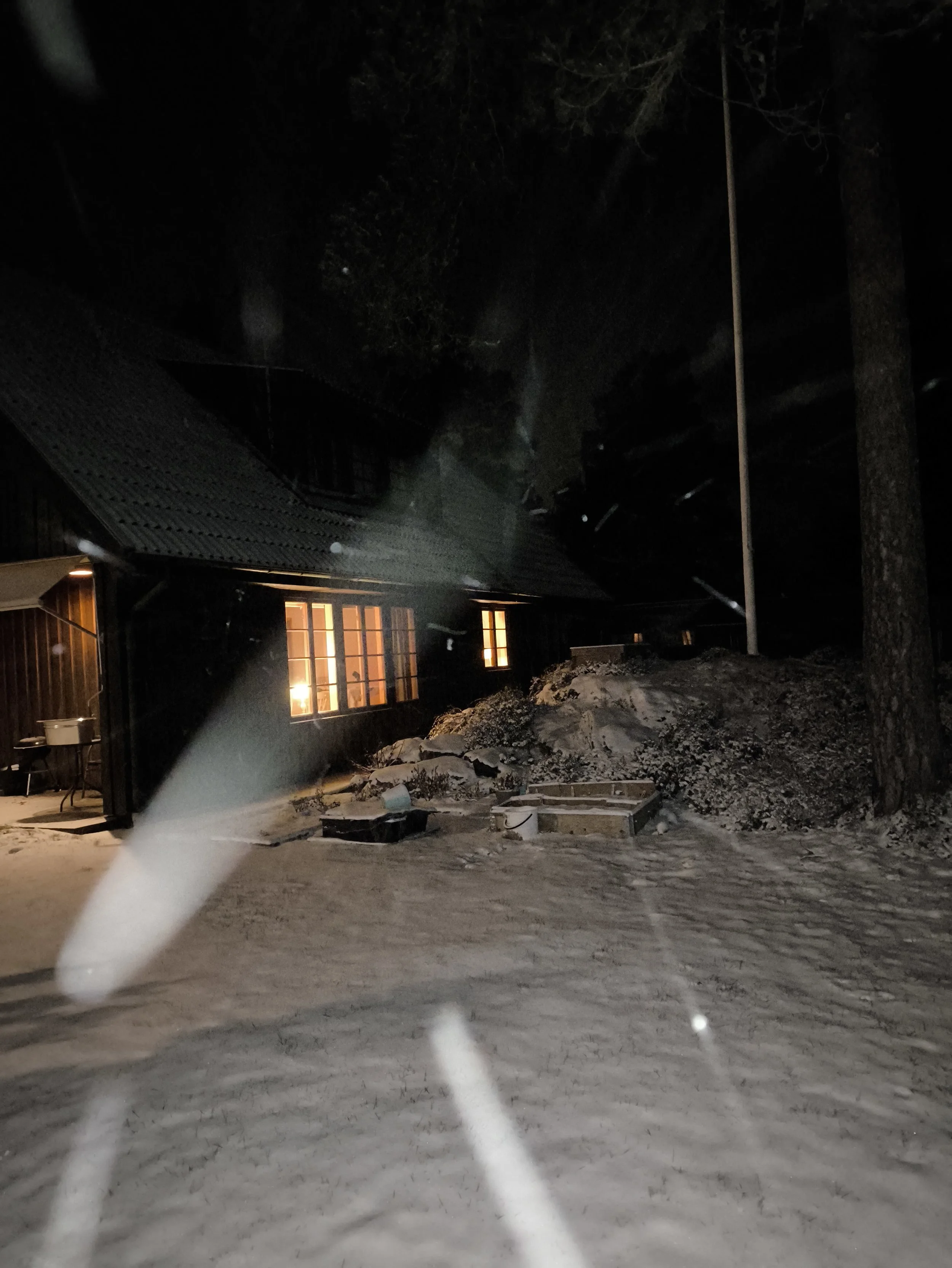 A night scene outside a house with illuminated windows, a snowy ground, trees, a white pole, and some construction or landscaping materials. West Cottage Sweden, Åsensbruk