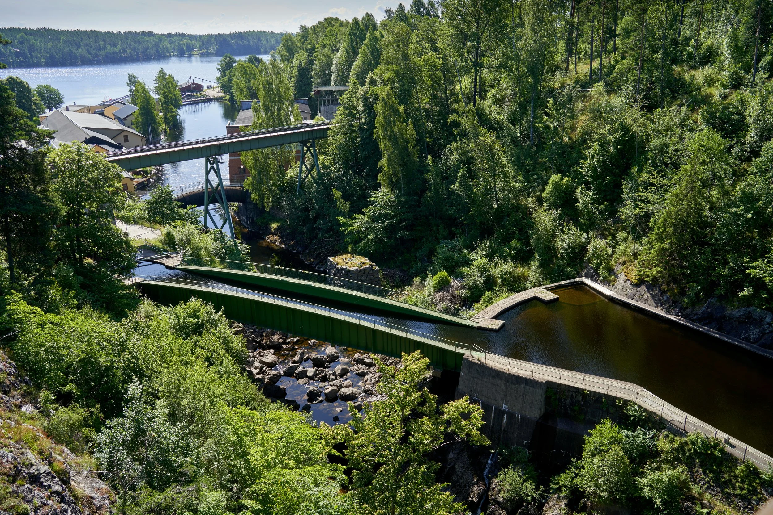 A scenic view of a lush green forest surrounding a river with multiple bridges, including a swing bridge and a dam, near a small town, with a lake in the background on a sunny day. Dalsland Canal.