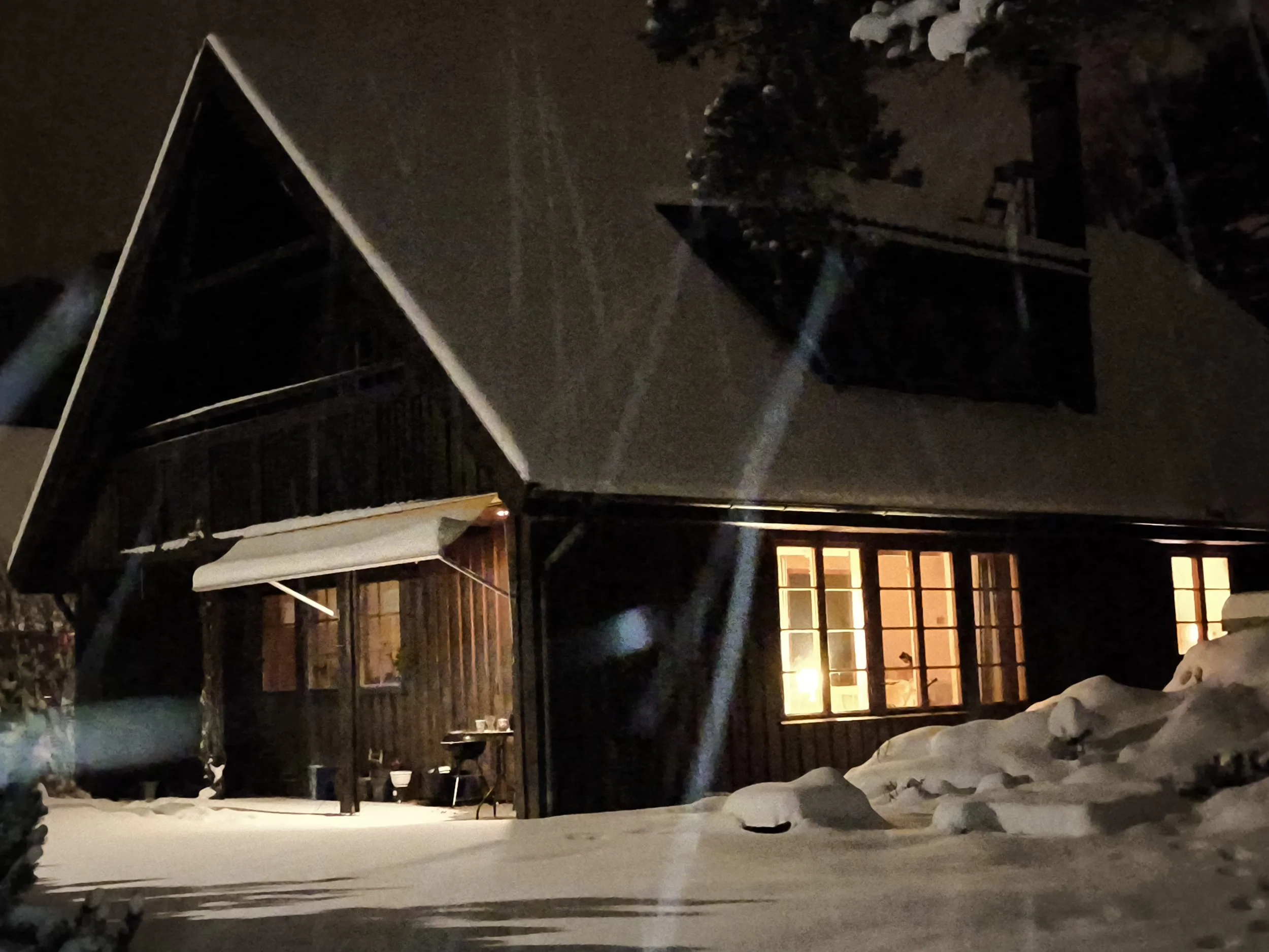 Nighttime scene of a snow-covered backyard with a raised wooden deck, a patio table, and chairs. Snow is accumulated on the ground, and there is a house with illuminated windows in the background. Tall trees are also visible. West Cottage, Åsensbruk