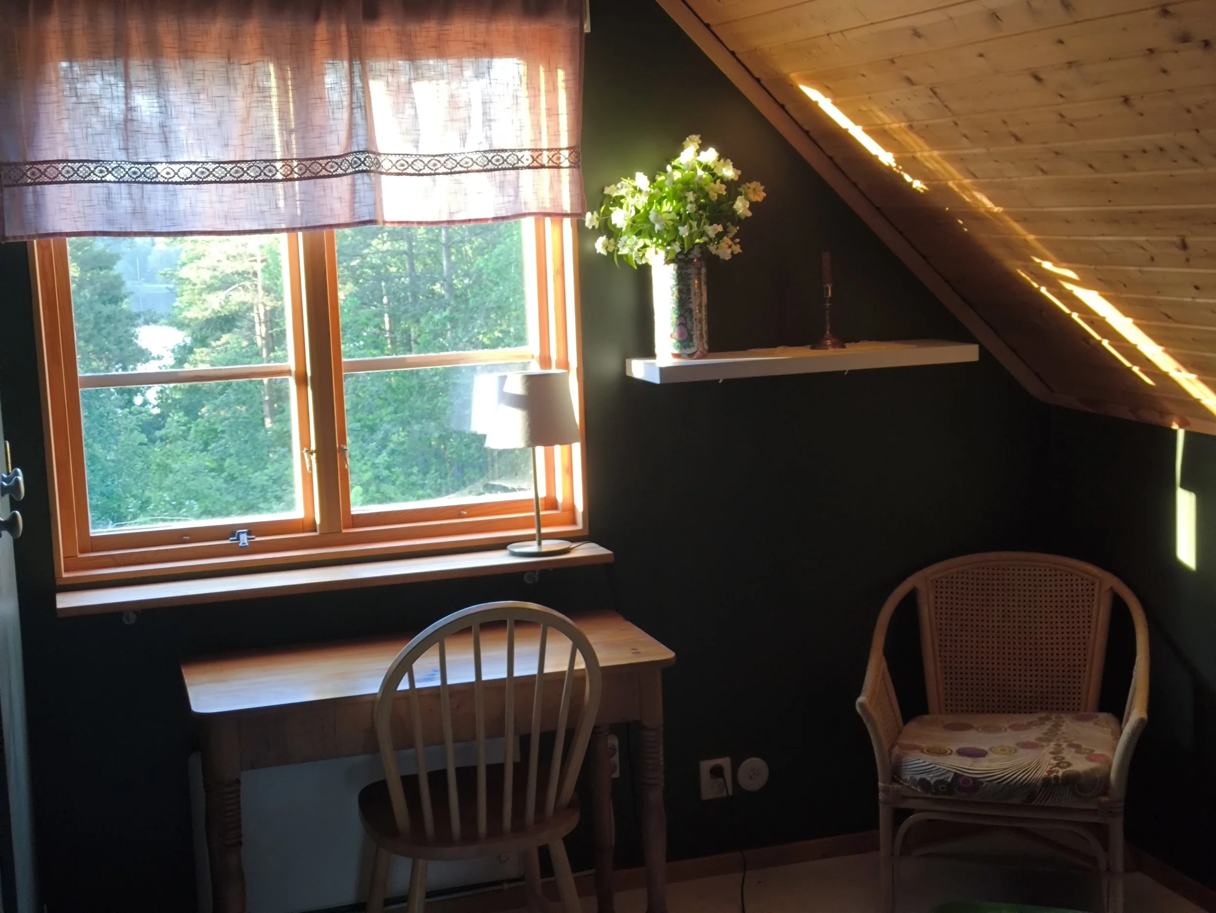 A cozy attic room with a large window overlooking a green, leafy outdoor scene, a small white shelf with a vase of white flowers, a table lamp, and three chairs of different styles and colors.