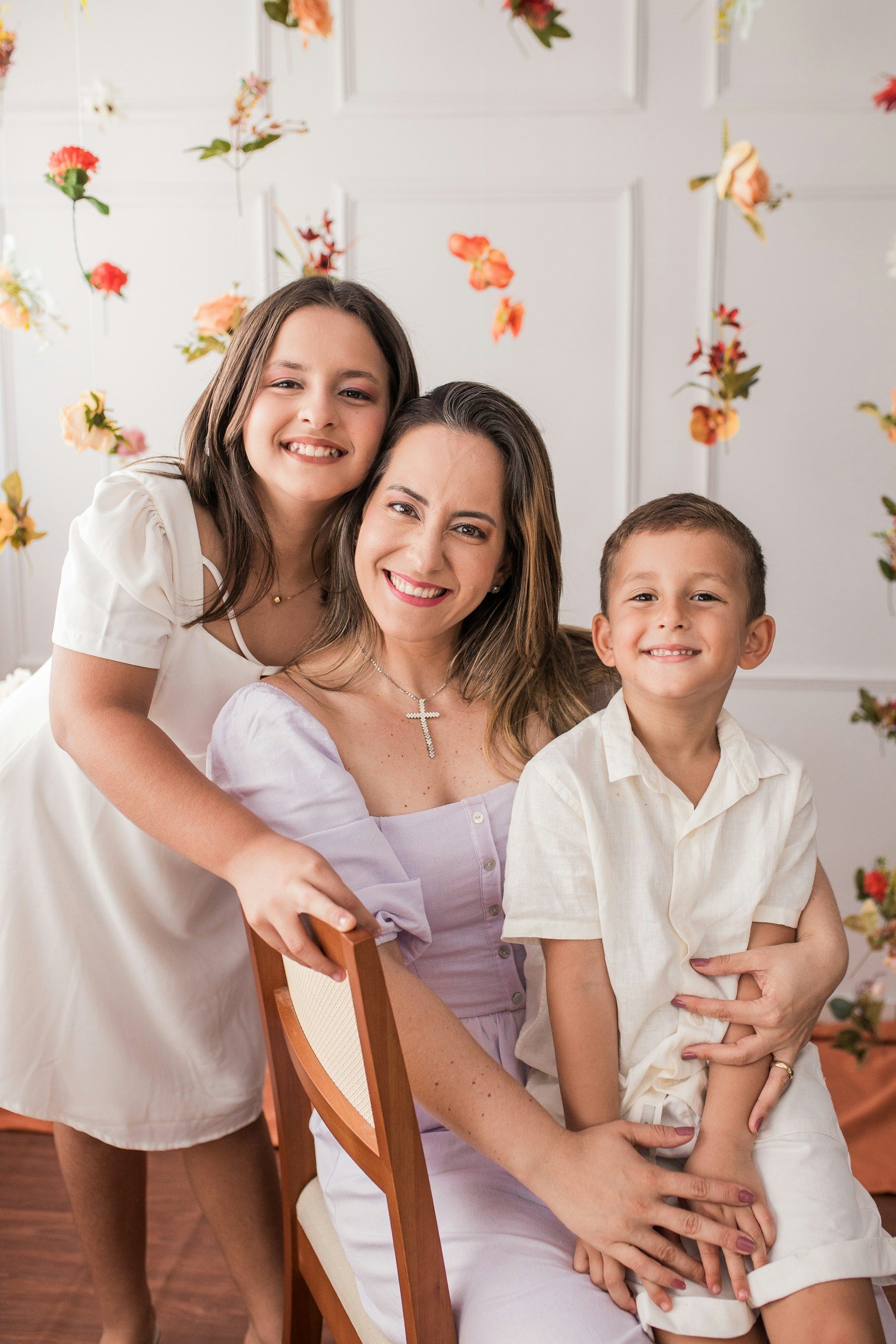 A woman sitting on a chair with a young girl and a young boy, all smiling and posing indoors with a background decorated with colorful flowers.