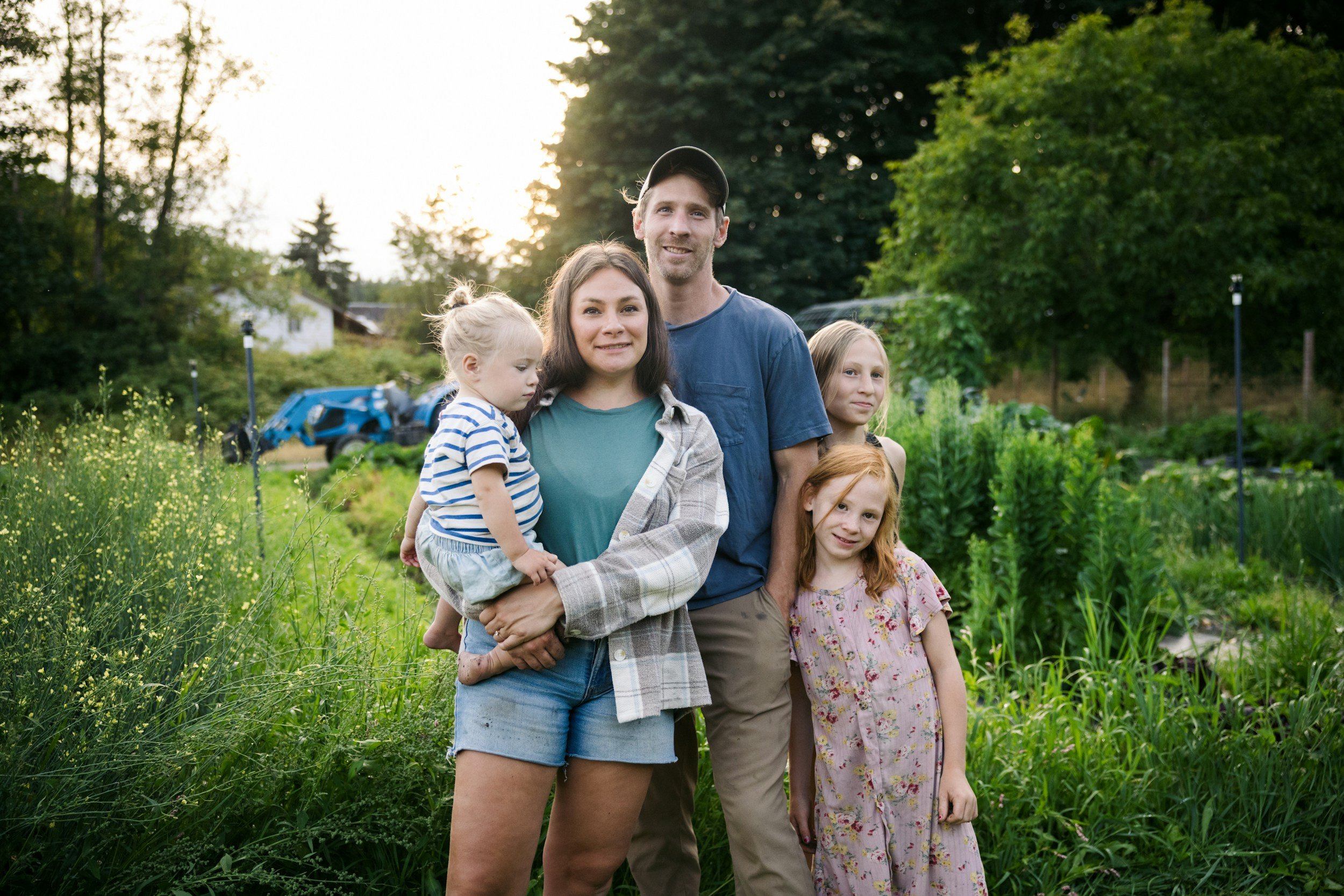 A family of five, including two children, standing outdoors in a garden or farm, with trees and sunlight in the background.
