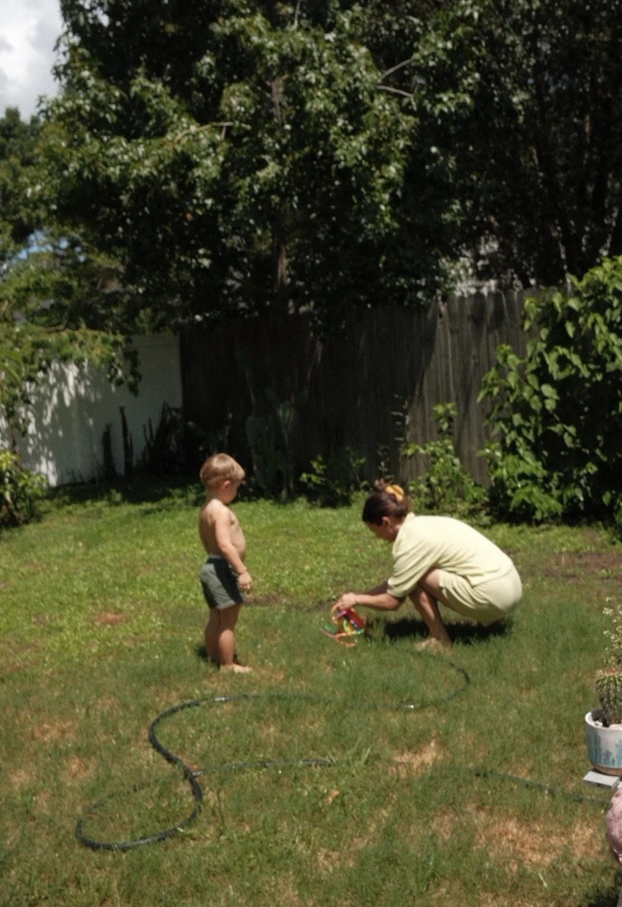 A woman crouching down in a backyard with a young boy standing nearby, both looking at a colorful toy. The scene is set on a grassy area with trees and a wooden fence in the background.