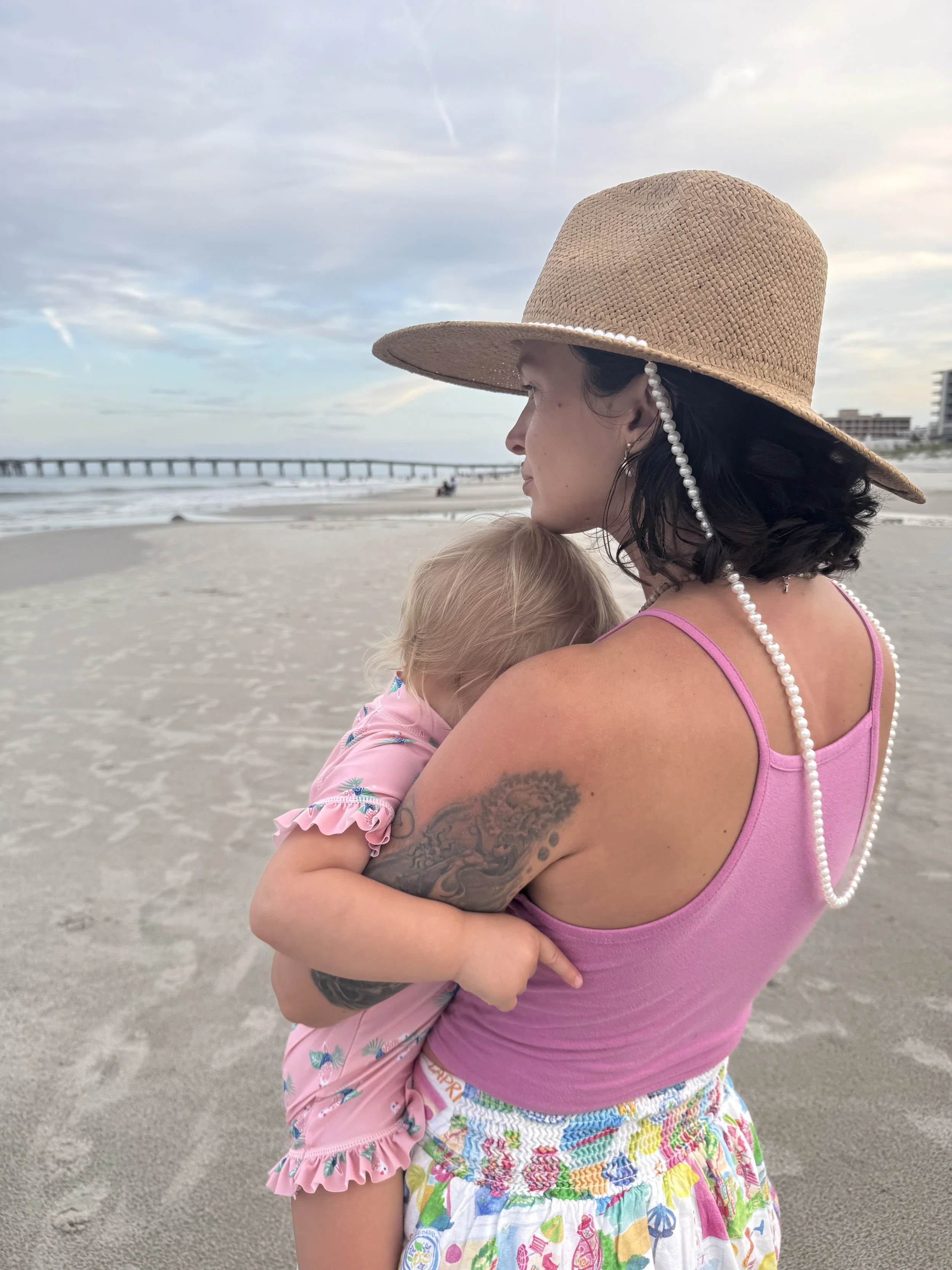 A woman wearing a wide-brimmed straw hat with a string of beads, holding a young girl in her arms, on a sandy beach with the ocean and pier in the background.
