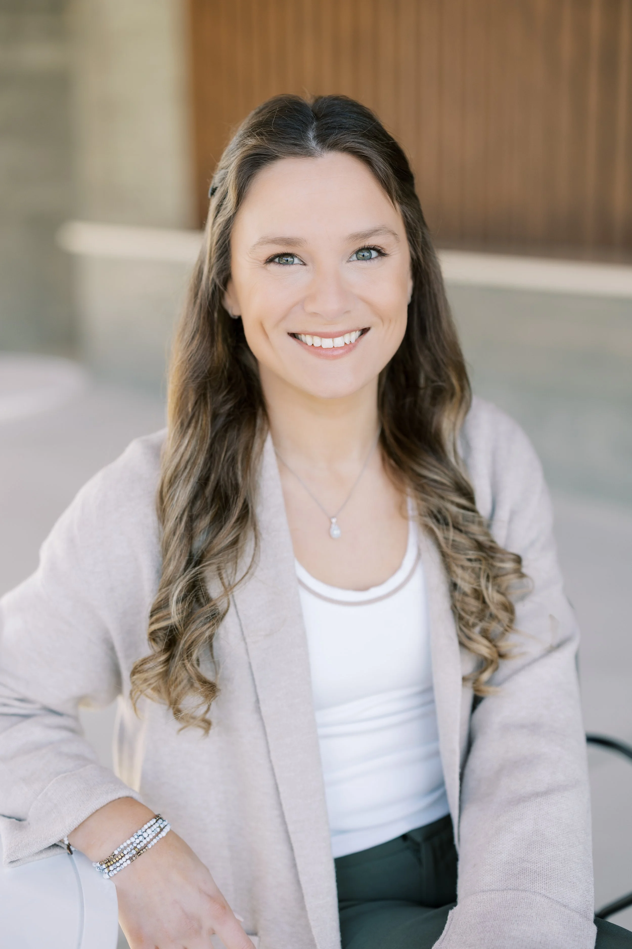 A smiling young woman with long, wavy blonde hair, wearing a white t-shirt and blue jeans, standing in front of a large window with black frames.