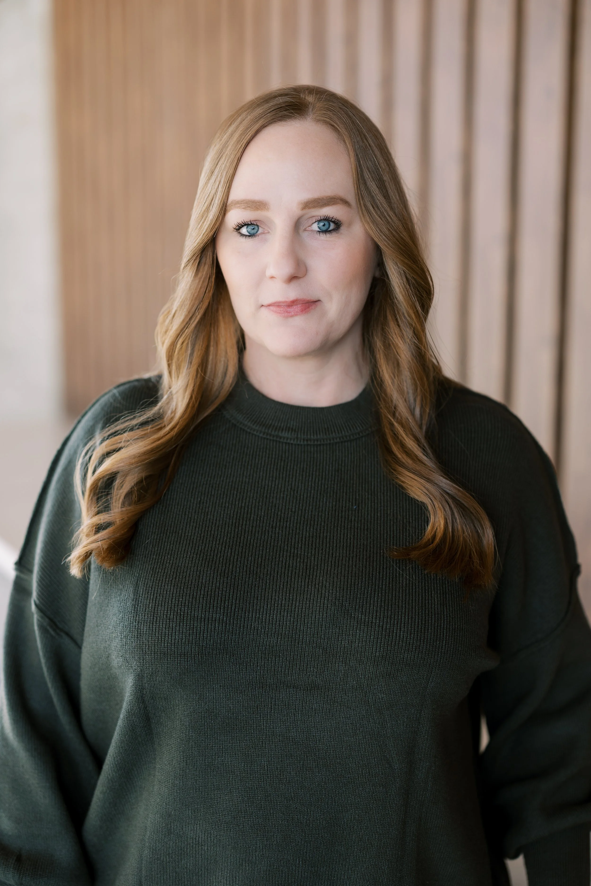A woman with long red hair and a green long-sleeve shirt smiling and standing with her hand on her hip in an office space.