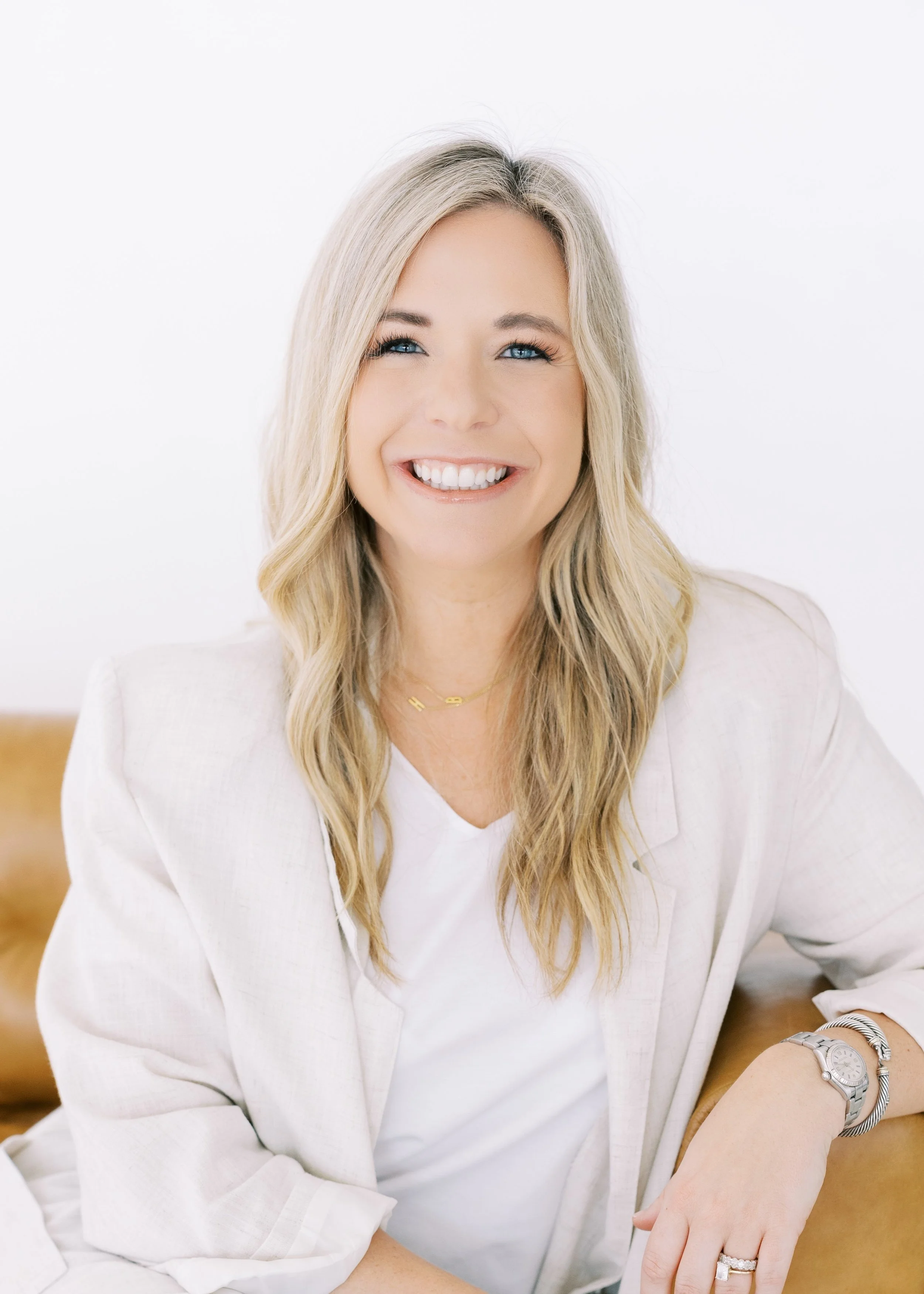 A woman with blonde hair and blue eyes smiling, sitting on a tan chair, against a pink and gray wall background.