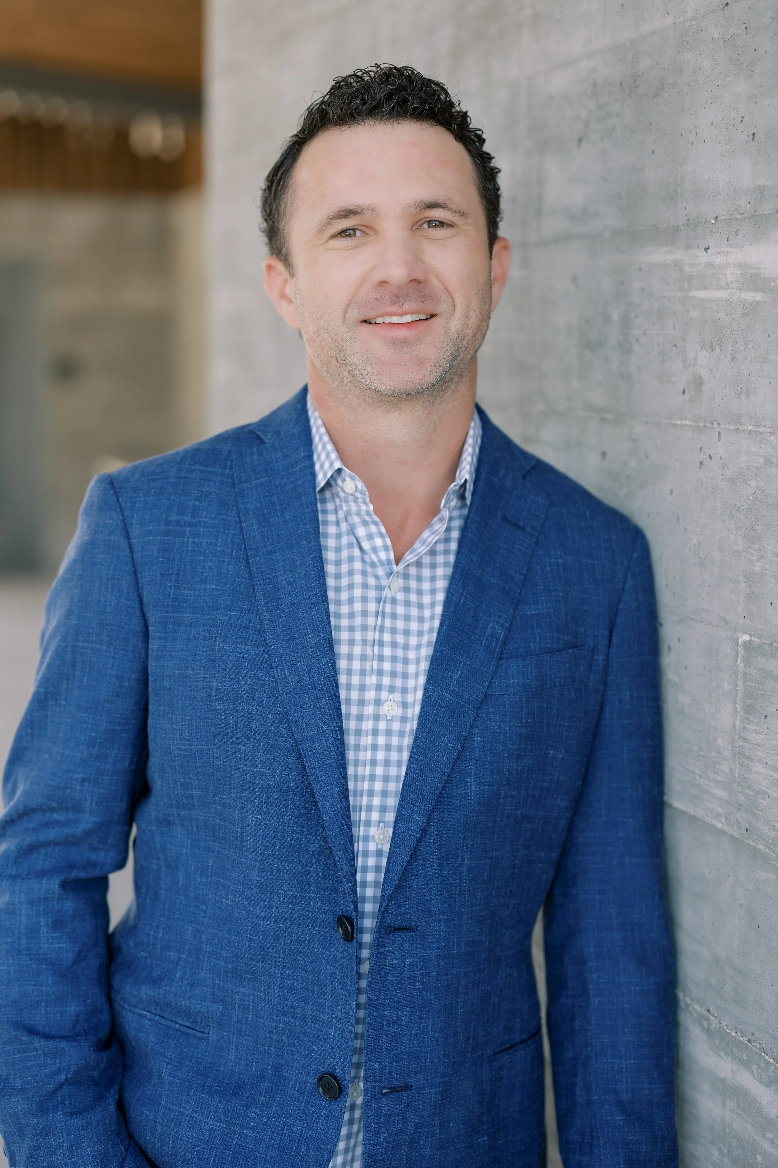 A man in a blue blazer and checkered shirt sitting outdoors in front of a modern building wall with greenery.