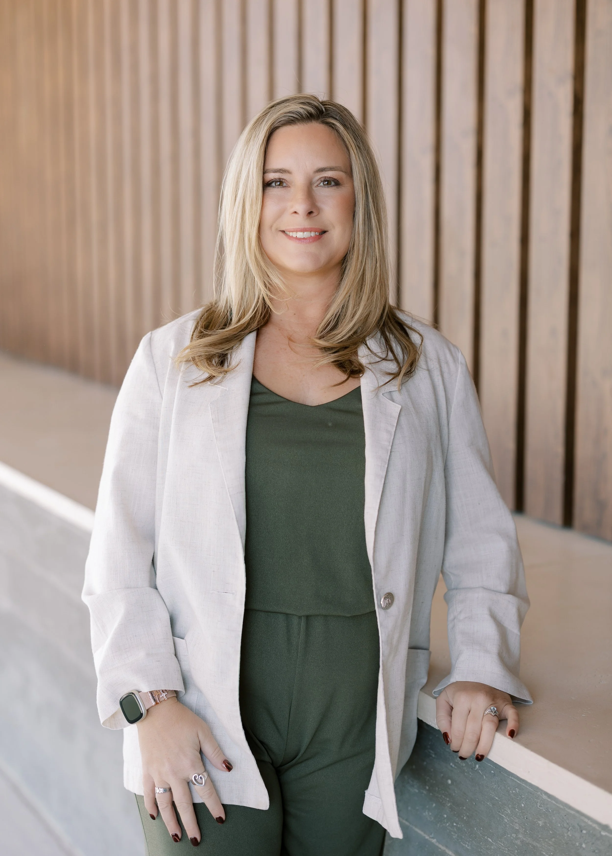 A smiling woman with shoulder-length blonde hair, wearing a cream-colored sweater with cold shoulder cutouts, sitting on a leather couch in front of a large window in a bright room.