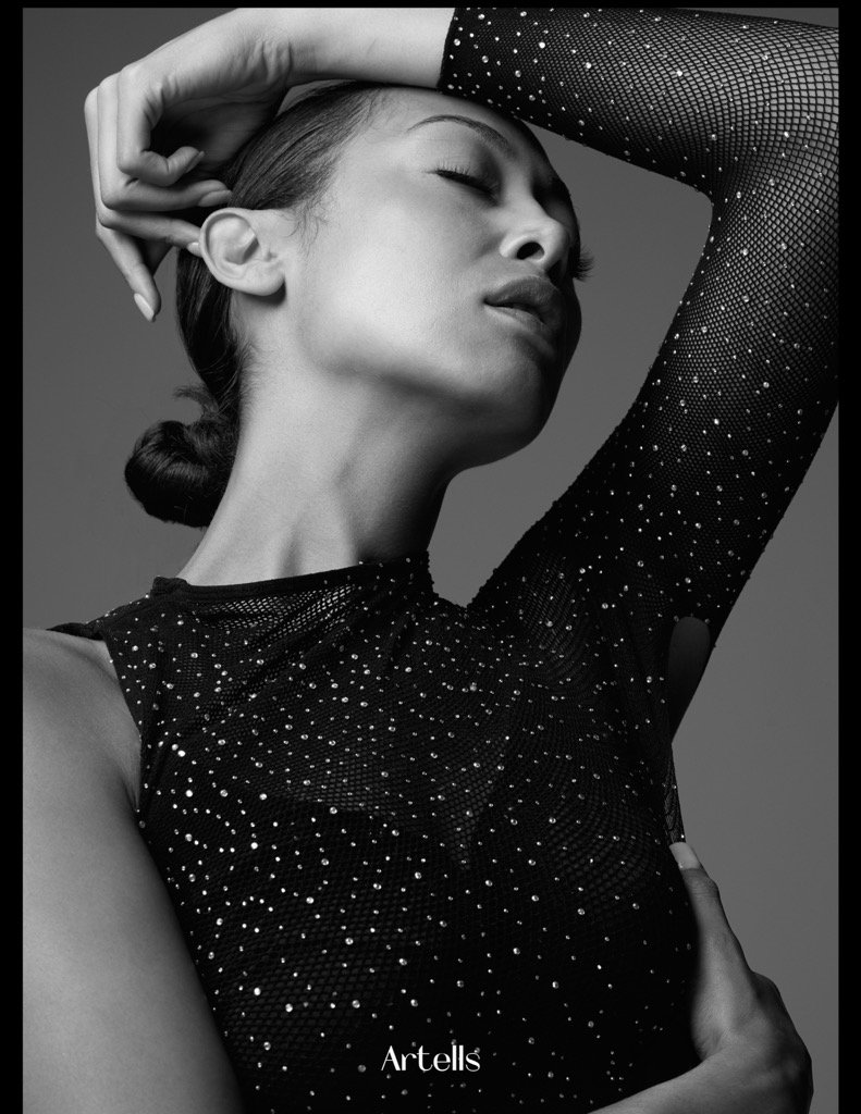 A stylish woman with her eyes closed, wearing a black, sheer, long-sleeved top embellished with small, sparkling dots, posing with one hand on her head and the other on her waist in a fashion photoshoot setting.