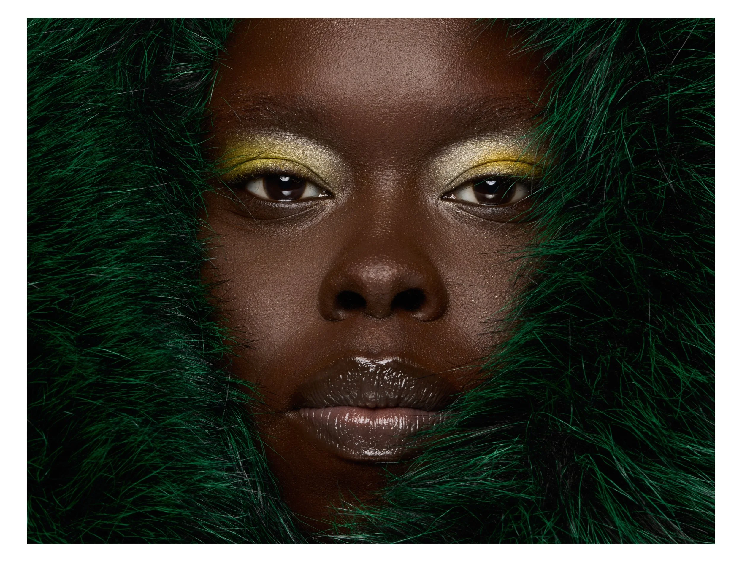 Close-up of a woman with dark skin, gold eyeshadow, and lips, framed by green fur.