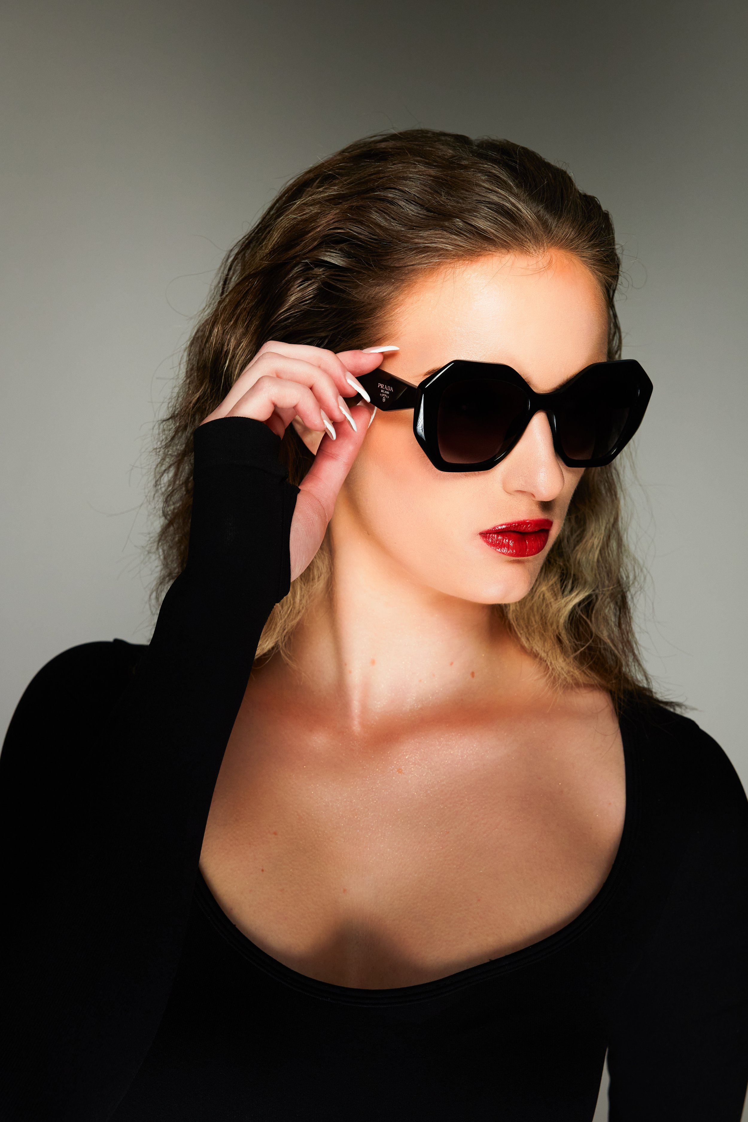 Close-up of a woman with fair skin, brown hair, blue eyes, wearing a black top, touching her face with her left hand, which has a small red heart tattoo on her wrist, and a small mole above her lip.