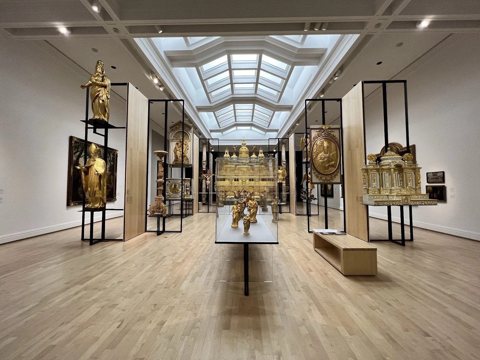 Une salle d'exposition avec des sculptures et objets en or, encadrés par des panneaux et une cage en verre, sous un plafond en verre.