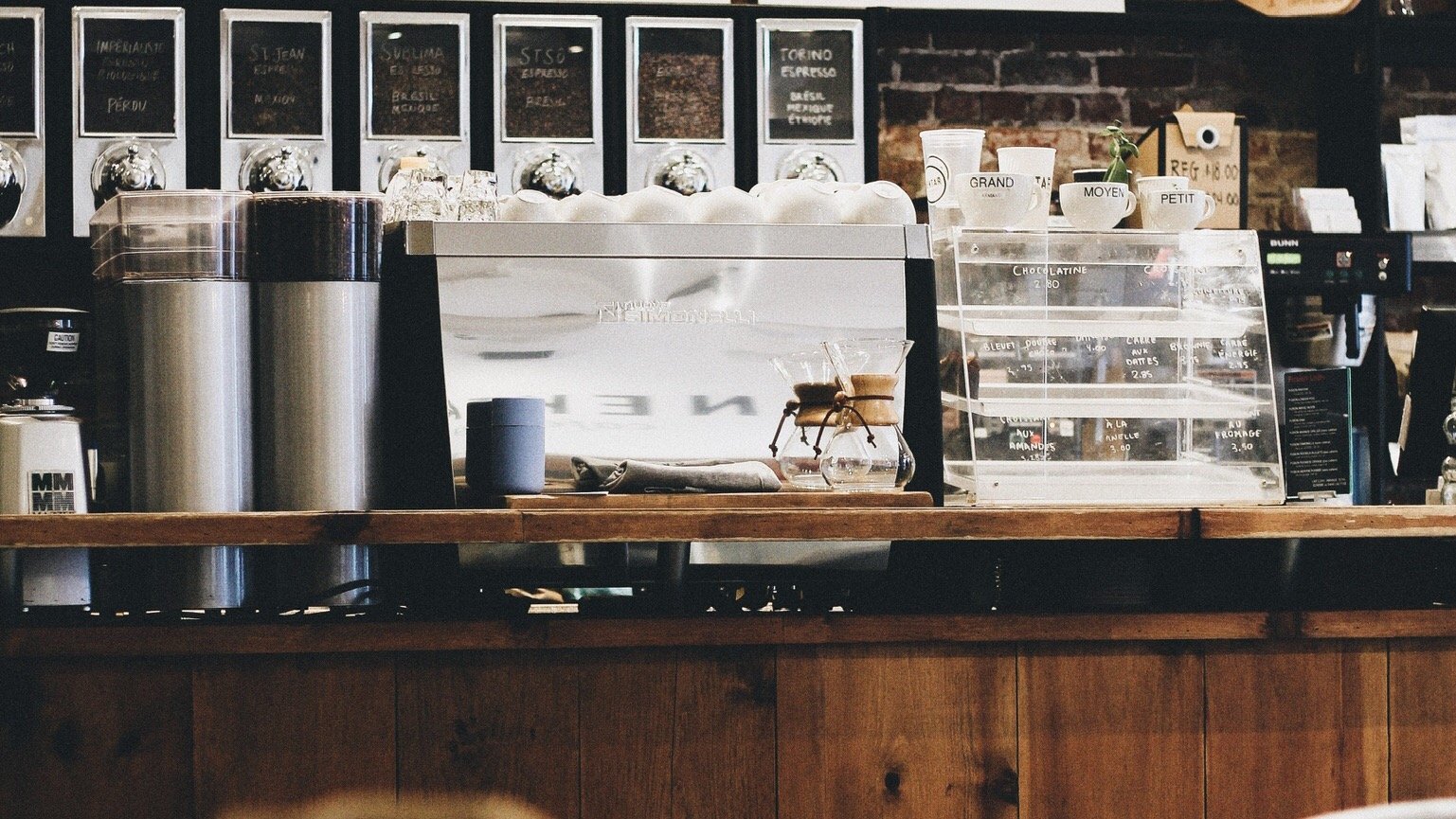 Comptoir de café avec machine à espresso, pots de sucres, tasses en céramique, et présentoir pour produits, décor en briques et bois.