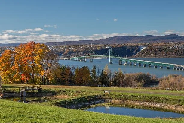 Paysage avec un pont suspendu au-dessus d'une rivière, arbres aux couleurs d'automne, pelouse et étangs, avec des montagnes en arrière-plan.