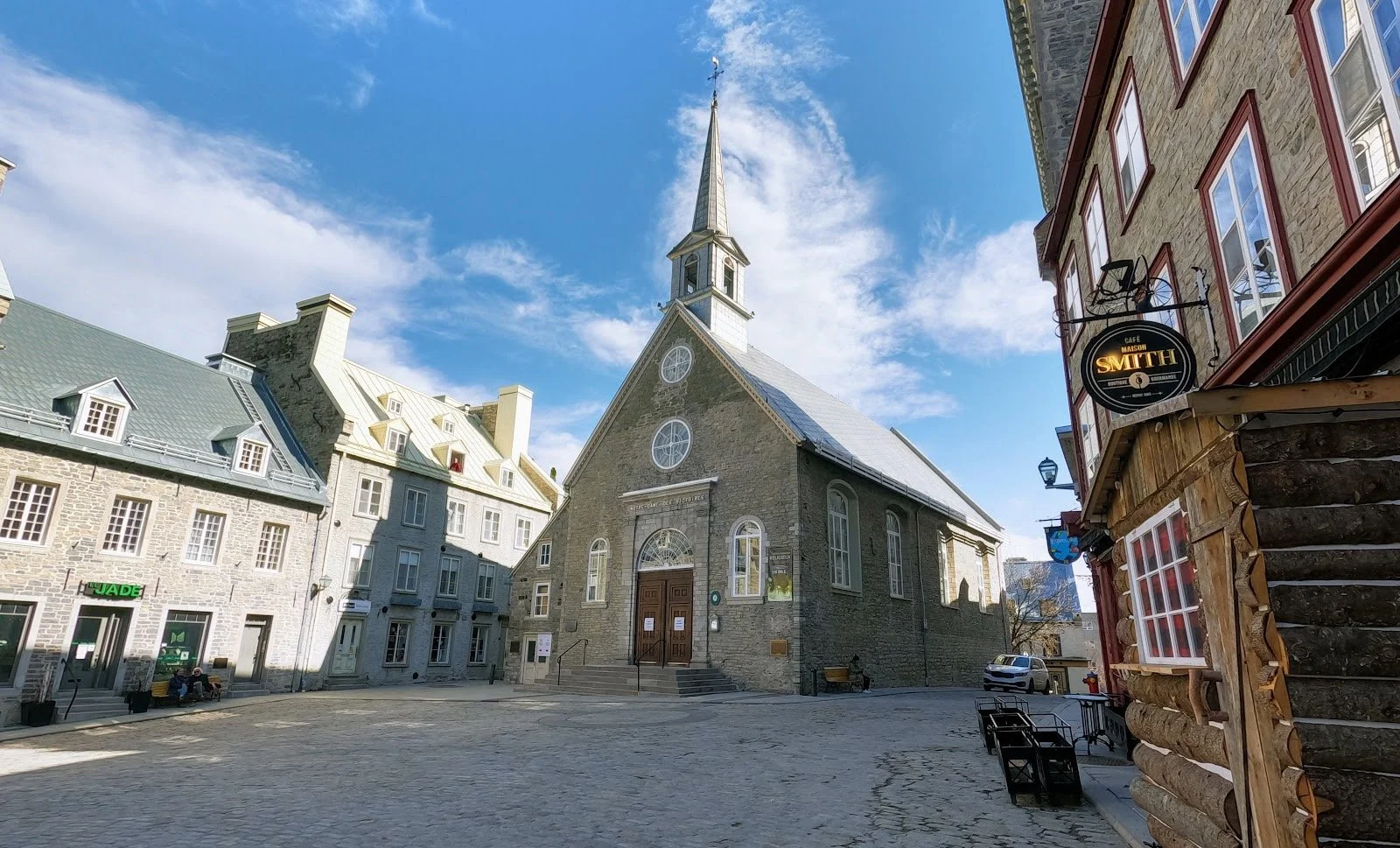 Une église en pierre avec un clocher, située au centre d'une place pavée, entourée de bâtiments anciens en pierre et en bois, sous un ciel bleu avec quelques nuages.