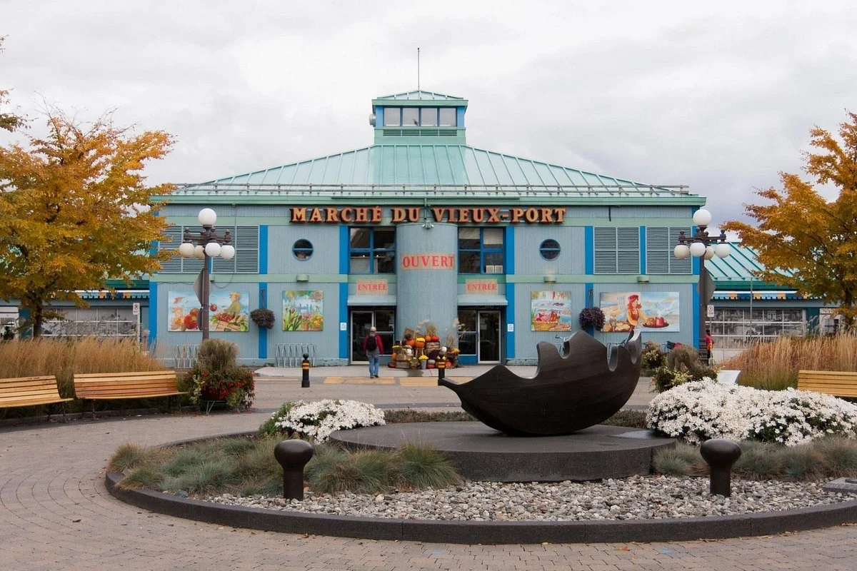 Un bâtiment bleu nommé 'Marché du Vieux-Port' avec des affiches de rue, entouré d'arbres d'automne, de bancs en bois, de lampadaires et d'une sculpture en forme de coquillage en métal noir au centre d'une zone piétonne
