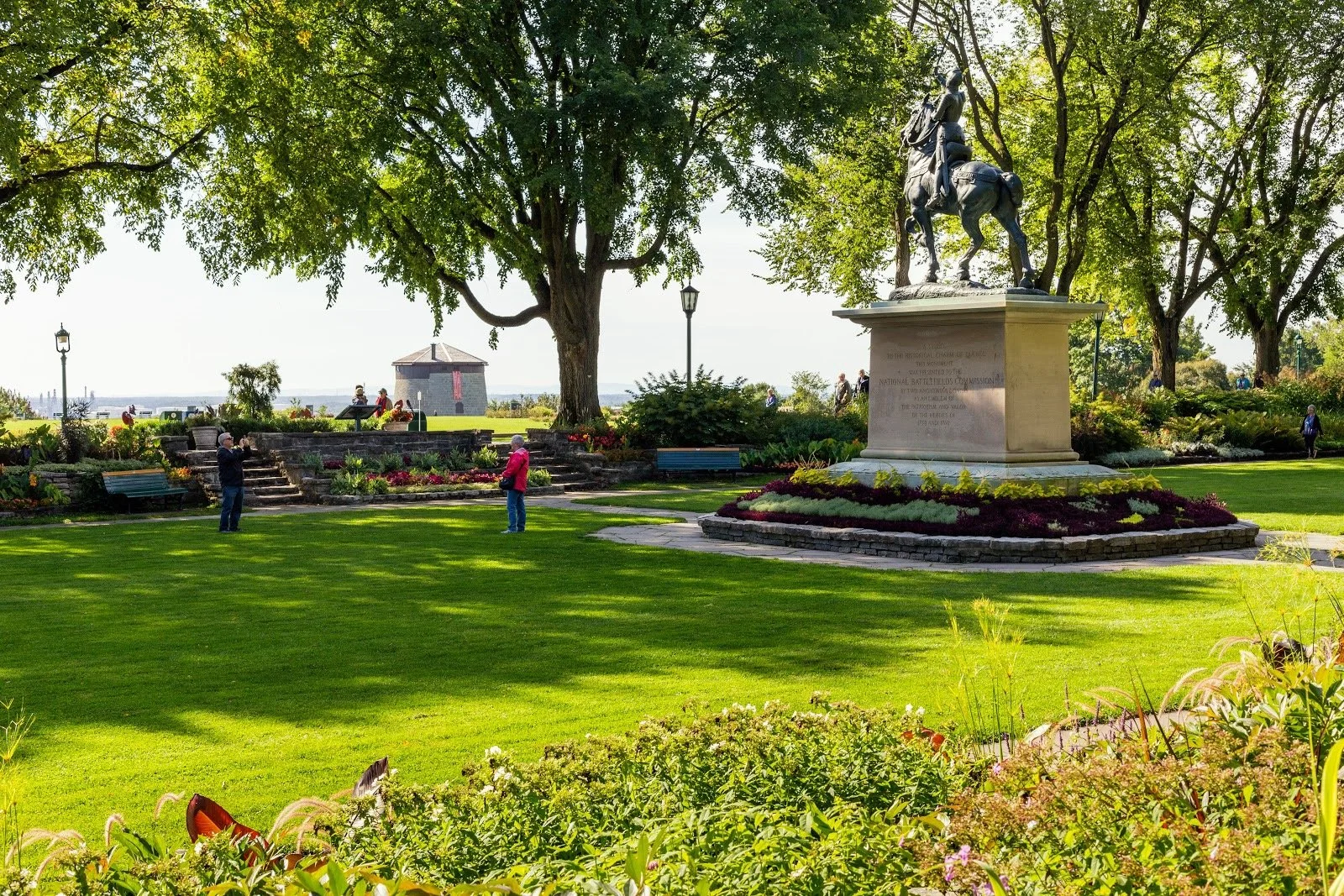 Parc en plein air avec une statue équestre et plusieurs personnes se promenant et prenant des photos, entouré d'arbres et de fleurs, ensoleillé.