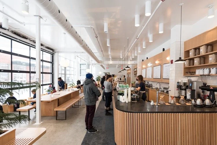 Intérieur d'un café moderne avec des clients ligne au comptoir, grandes fenêtres, décoration minimaliste en bois, et personnes assises à des tables, ambiance lumineuse.