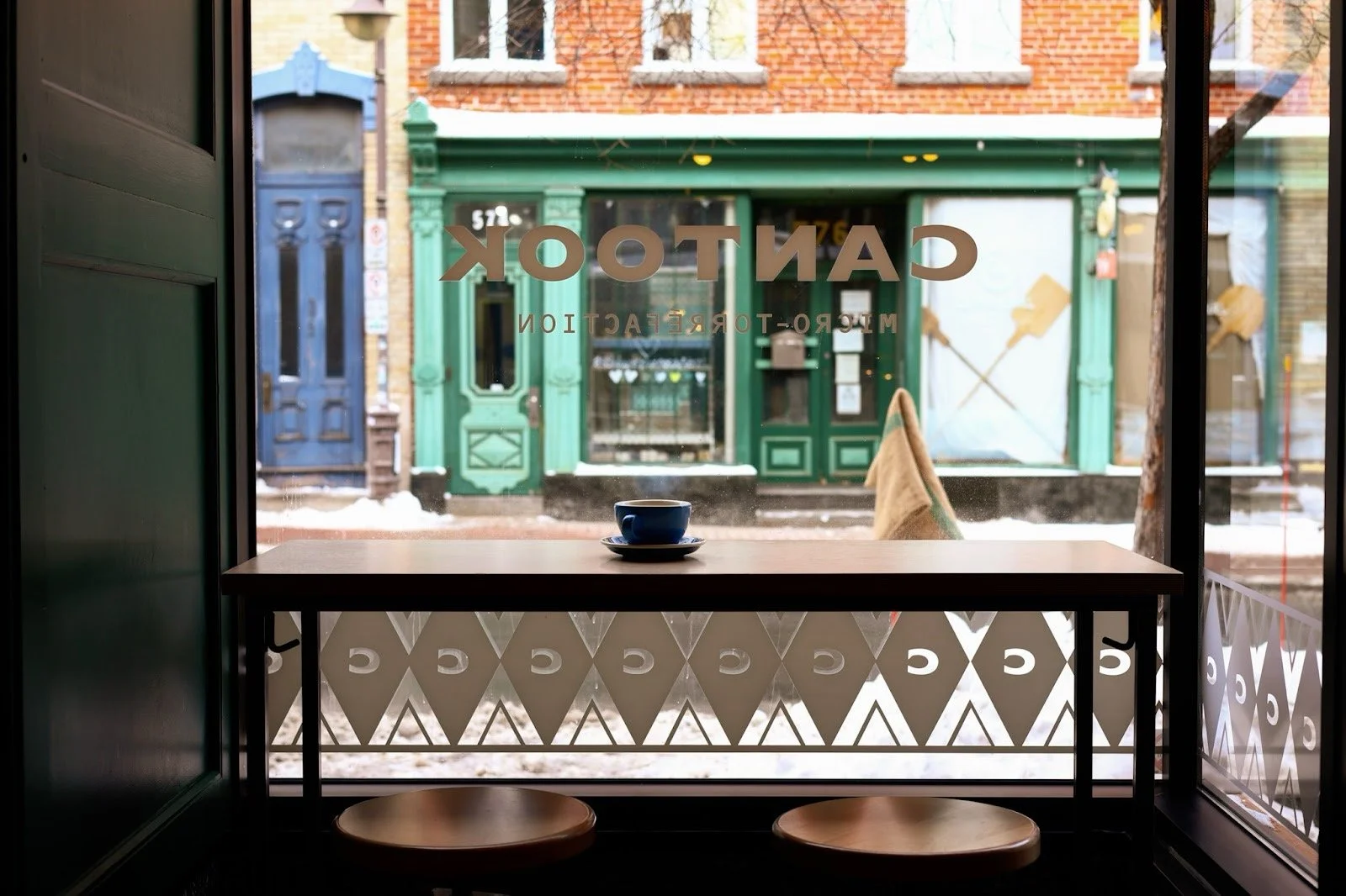 Vitrine d'une cafétéria avec une tasse de café sur une table devant une fenêtre montrant la rue avec des bâtiments en briques et une façade verte.