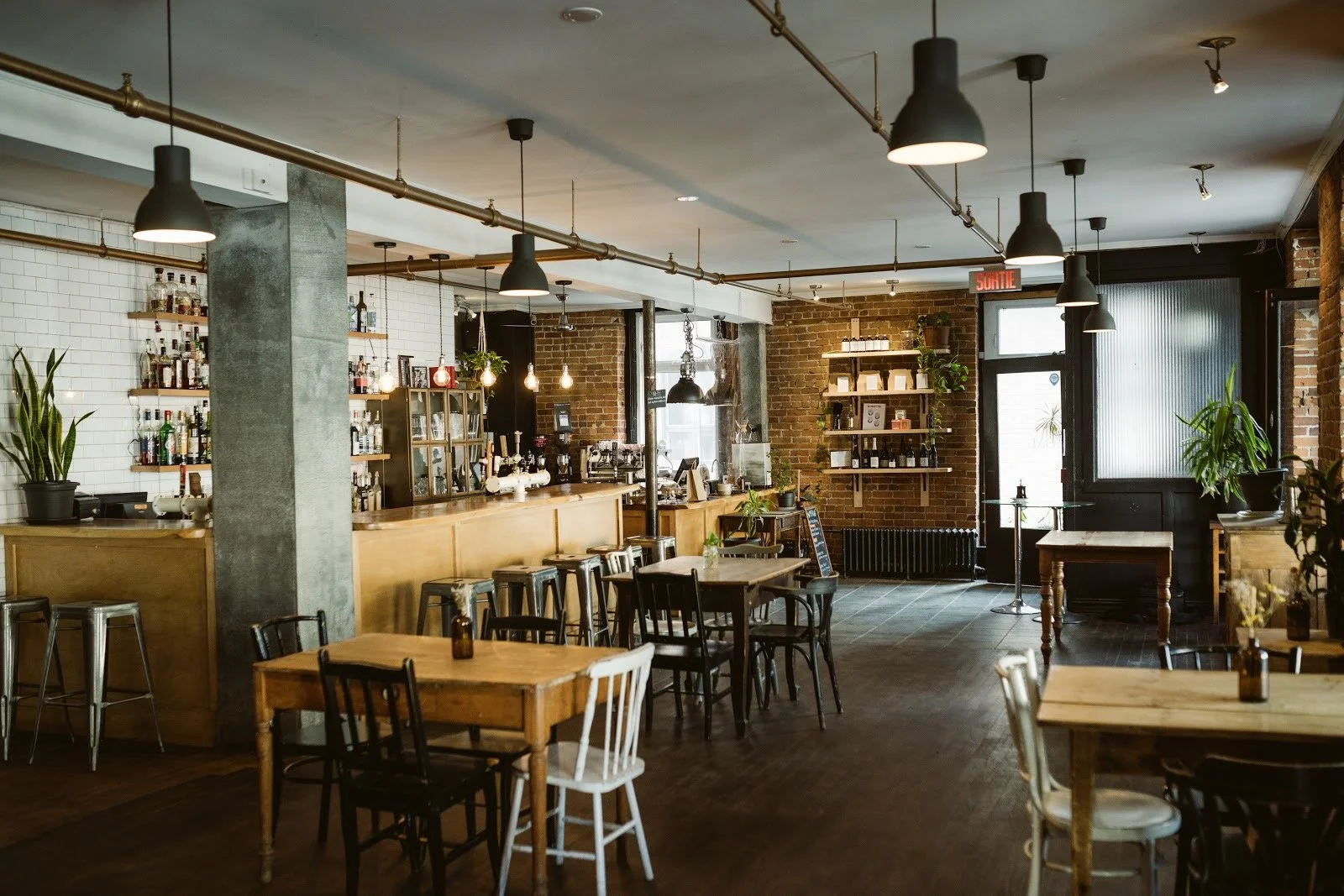 Intérieur d'un café ou bar avec comptoir en bois, étagères de bouteilles, tables en bois, chaises variées, mur en briques, plantes vertes, éclairage suspendu, porte de sortie avec panneau lumineux