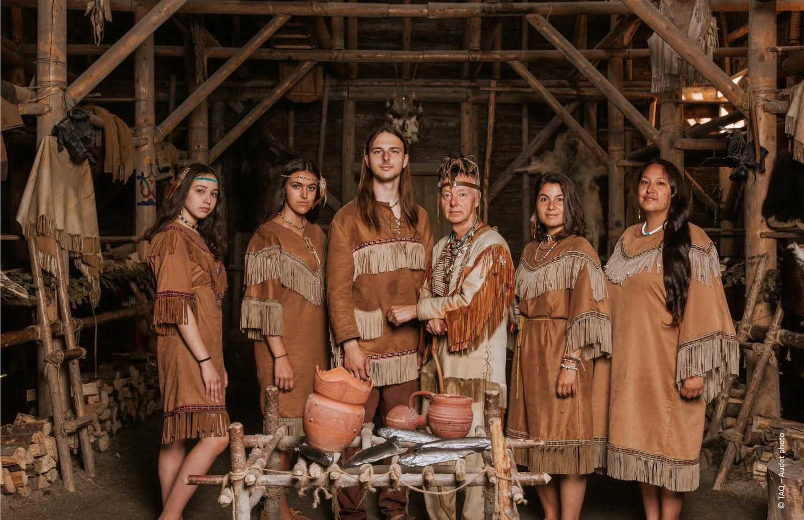 Groupe de personnes en costumes traditionnels amérindiens devant une structure en bois, avec des poteries en terre cuite et du poisson sur une table en bois.