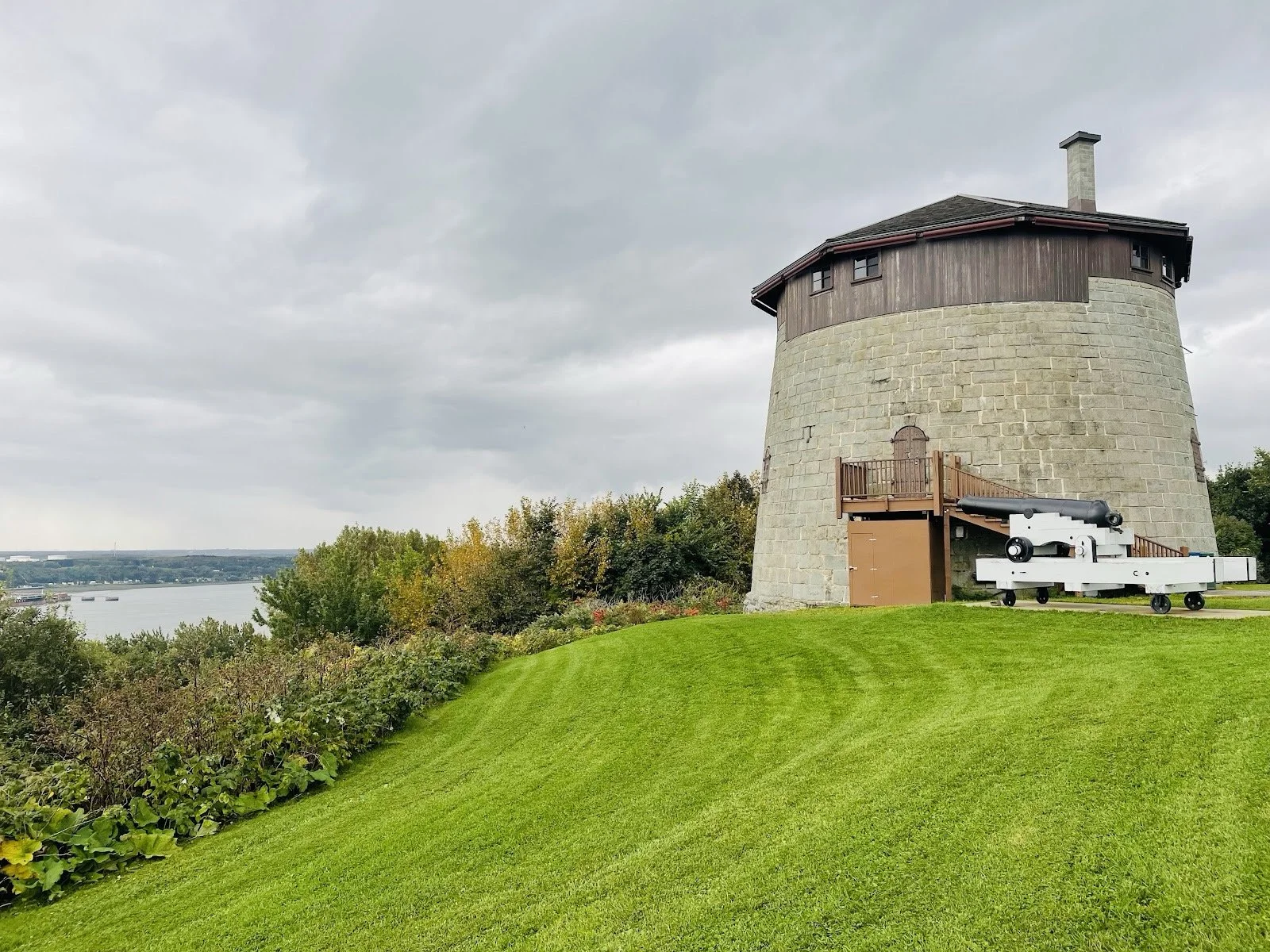 Tour de guet en pierre avec canon en position sur la pelouse verte, vue sur la rivière et la nature environnante sous un ciel nuageux.