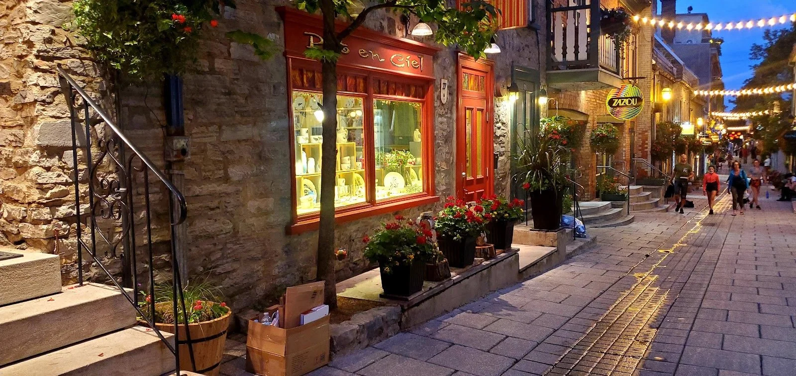 Rue piétonne éclairée par des lumières, avec une boutique de vaisselle et une terrasse de café, des fleurs en pot, et des personnes marchant au crépuscule.
