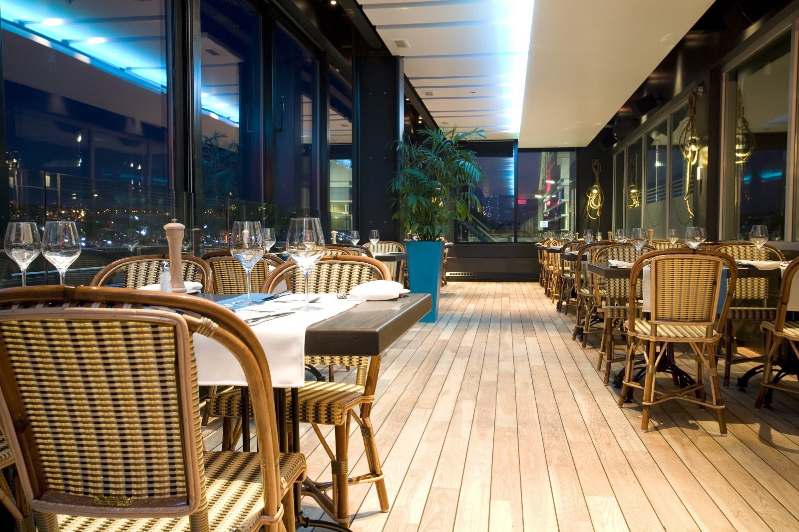 Salle à manger élégante avec des tables dressées, des chaises en rotin, des verres à vin, une grande plante en pot, et des grandes fenêtres offrant une vue de la ville la nuit.