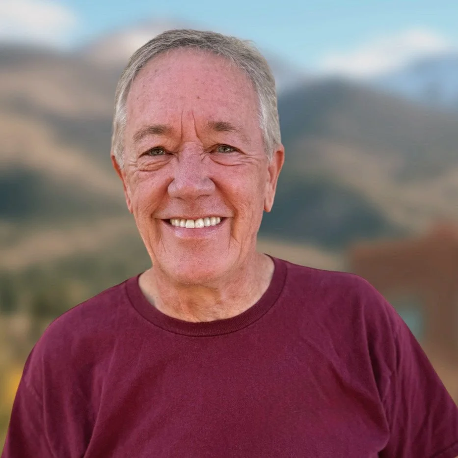 A smiling older man with salt and pepper hair and a beard, wearing a maroon shirt, outdoors with a background of mountains.