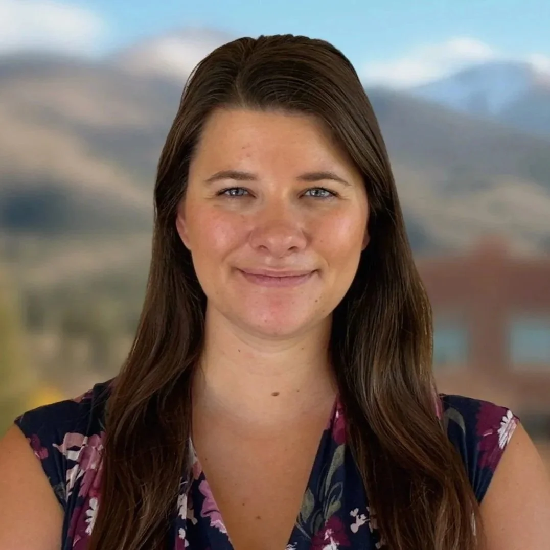 A woman with long brown hair and blue eyes smiling, standing outdoors with a backdrop of mountains, trees, and a building.