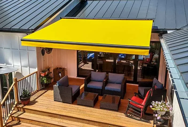 Image of a yellow awning covering a nice sitting area on a wood patio. The home has a dark metal roof.