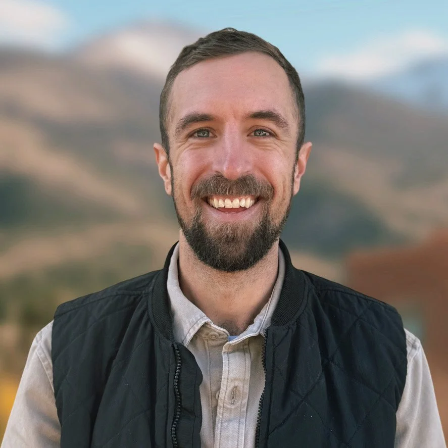 A smiling man with dark hair and a beard, wearing a black vest, with mountains in the background.