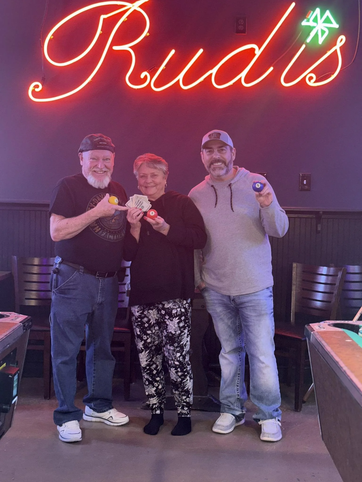 Three people with pool balls standing in front of a neon sign that says "Rudy's" inside a bar or game room.