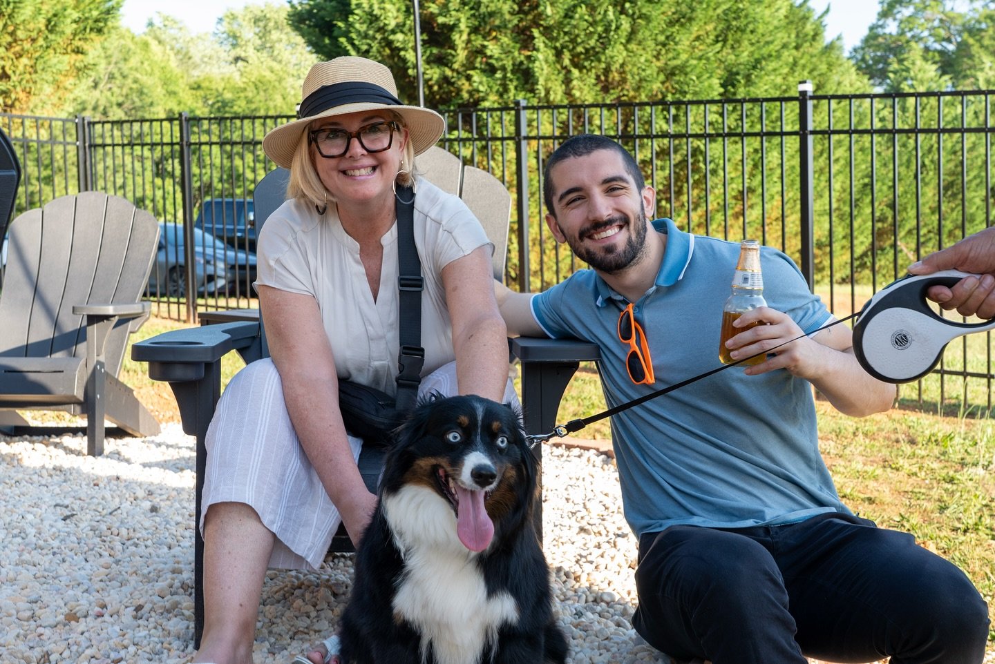 Two smiling people, a woman with glasses and a hat and a man with sunglasses, are sitting outdoors with a dog. The woman is sitting on a chair, and the man is kneeling beside her, holding a beer and a leash attached to the dog. The dog is sitting on 