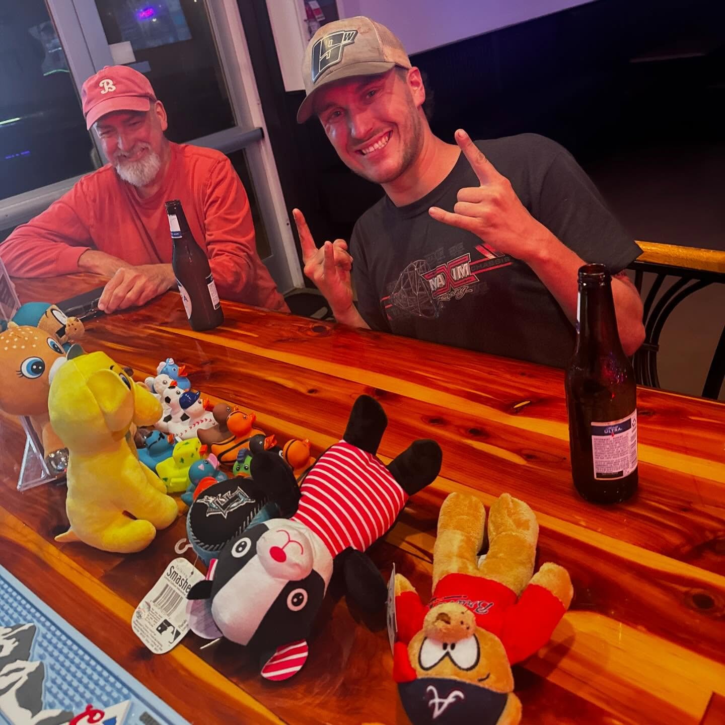 Two men sitting at a wooden table with plush toys and bottles of beer. One man is smiling, making a rock and roll gesture with his hands, wearing a gray cap and black T-shirt. The other man, older with a white beard and pink cap, is smiling and weari
