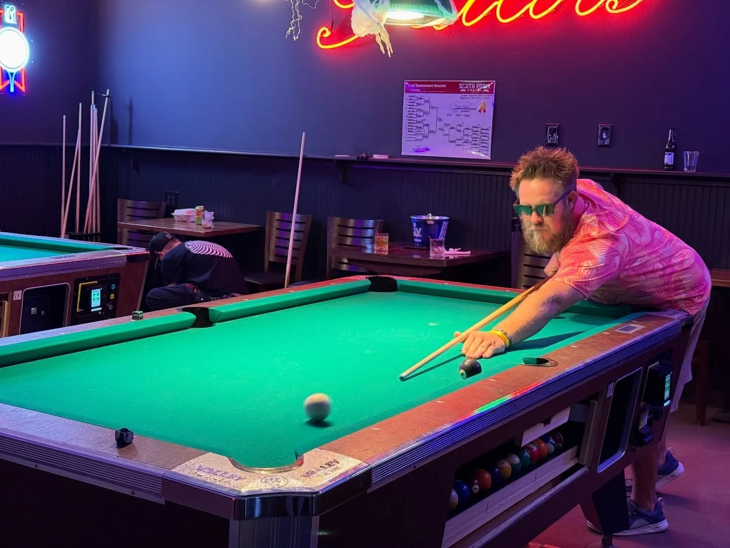 A man with sunglasses and a pink patterned shirt playing pool on a green felt table in a dimly lit bar with neon signs and tables in the background.