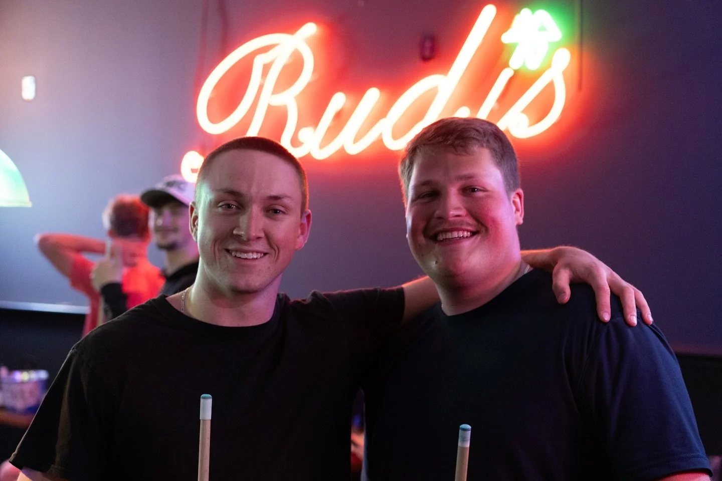 Two men smiling, with one having his arm around the other, in front of a neon sign that reads 'Bud's' in a bar or club setting with other people in the background.