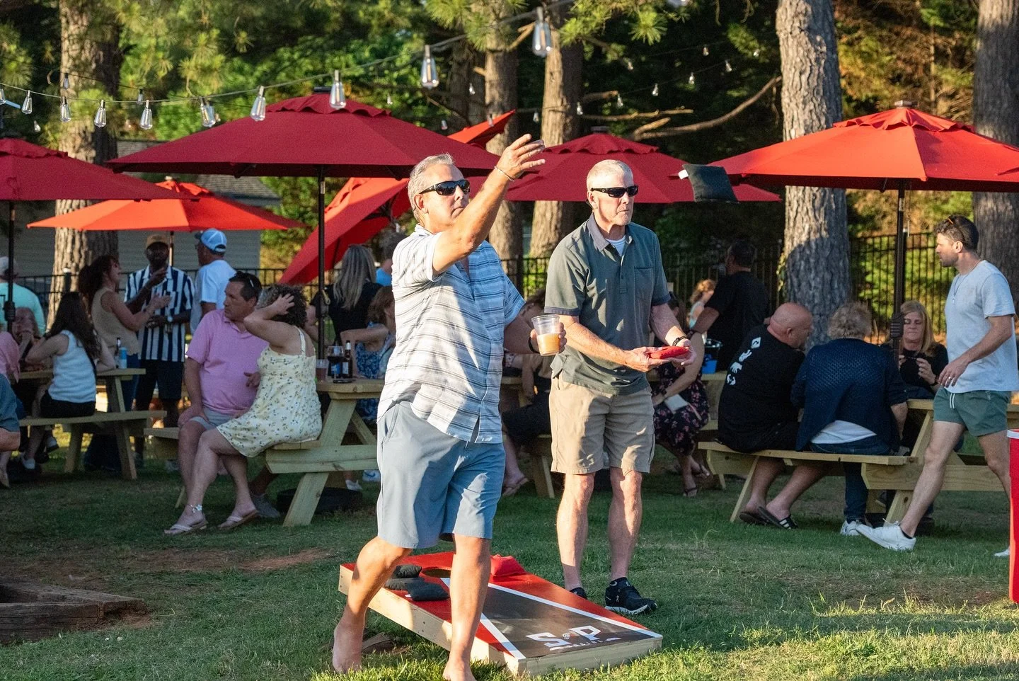 People playing cornhole at an outdoor gathering with picnic tables, red umbrellas, and string lights in a wooded area.