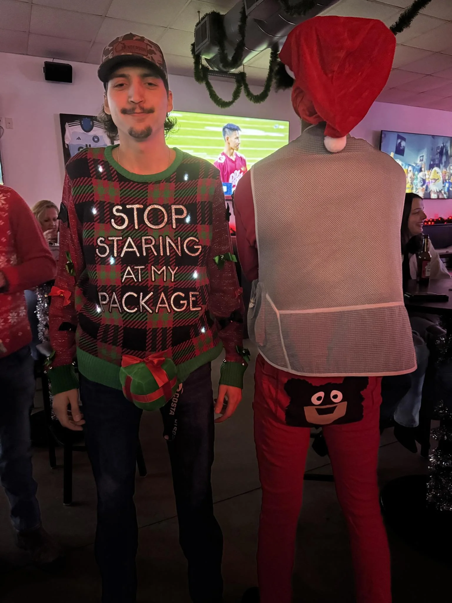Two people in Christmas-themed outfits at a party. The person on the left is a man wearing a Christmas sweater that says 'STOP STARING AT MY PACKAGE' with Christmas lights decorating it, and a Santa hat attached to his sweater. The person on the righ