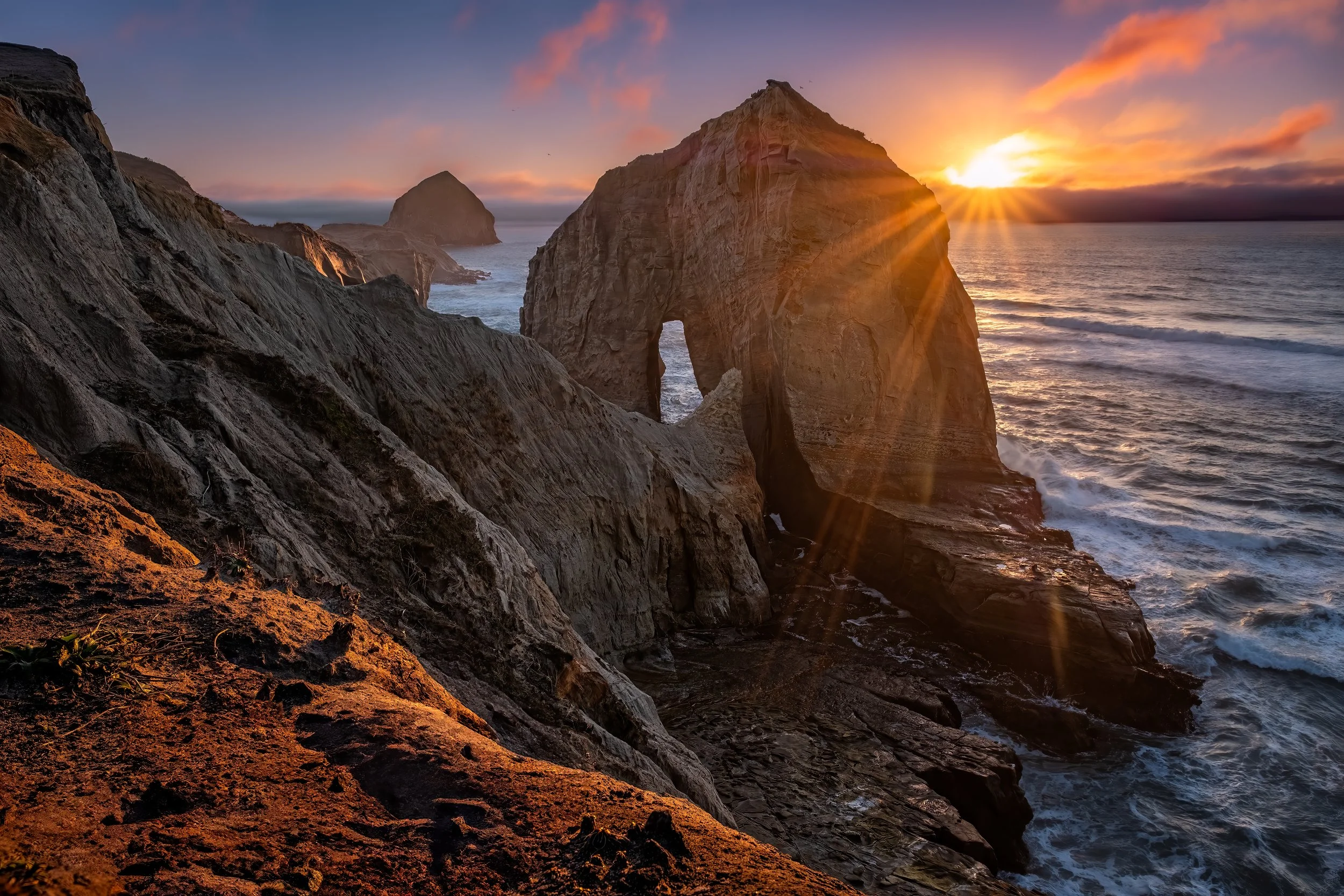 Sun setting behind large ocean cliffs with natural rock arch formation, waves crashing below, and colorful sky with clouds.