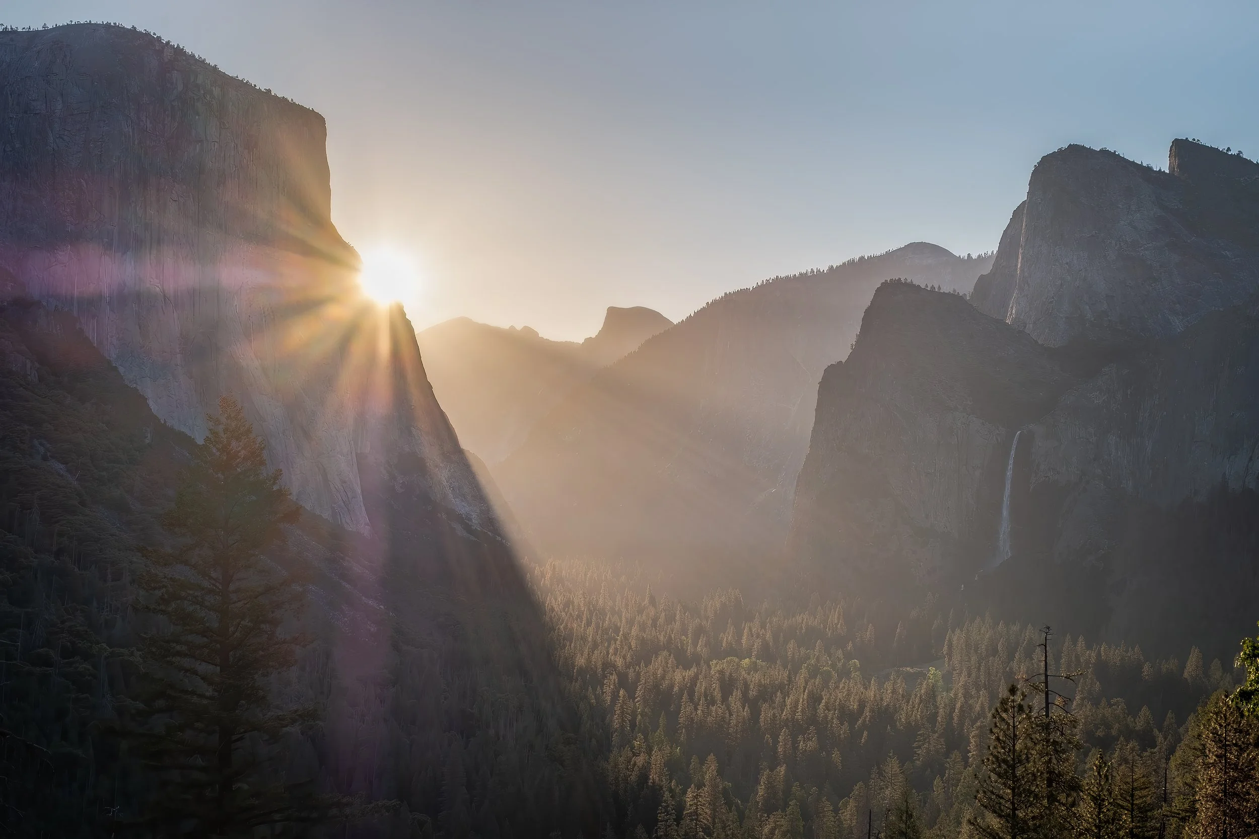 Sunrise over a valley with mountains and a waterfall, with sun rays illuminating the scene.