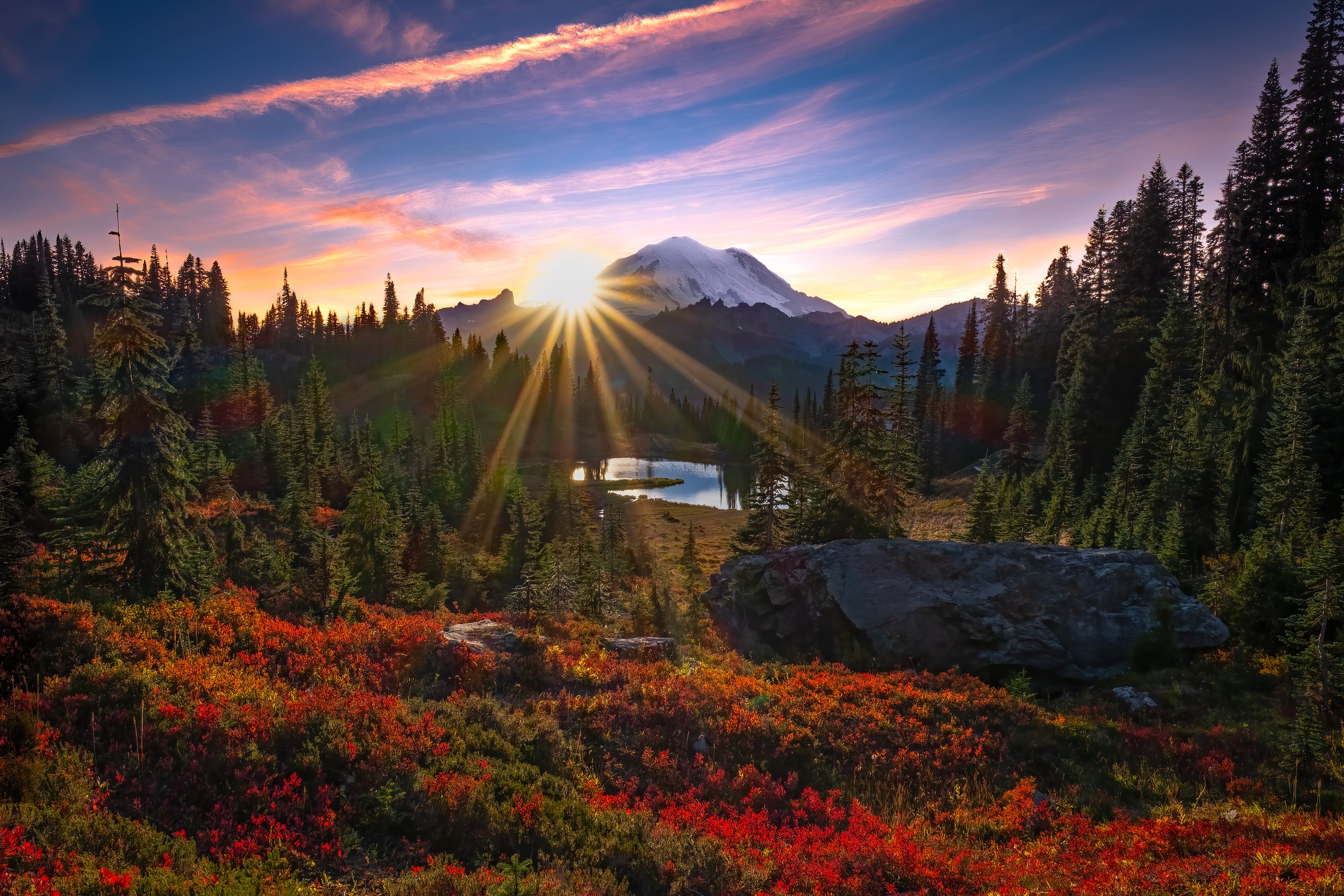 Sunrise over a snow-capped mountain with a forest and a pond in a valley, colorful autumn foliage in the foreground.