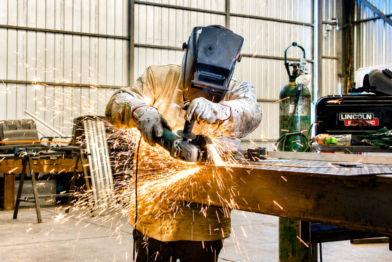 Worker welding metal in a workshop, wearing protective gear and welding helmet, with sparks flying.