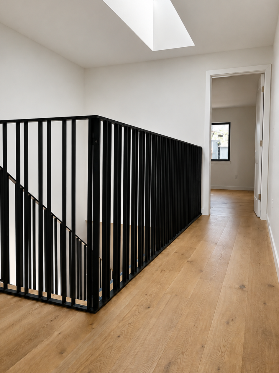 Interior view of a light-filled hallway with a staircase featuring a black metal railing, hardwood floors, a skylight, and an open doorway leading to a room with a window.