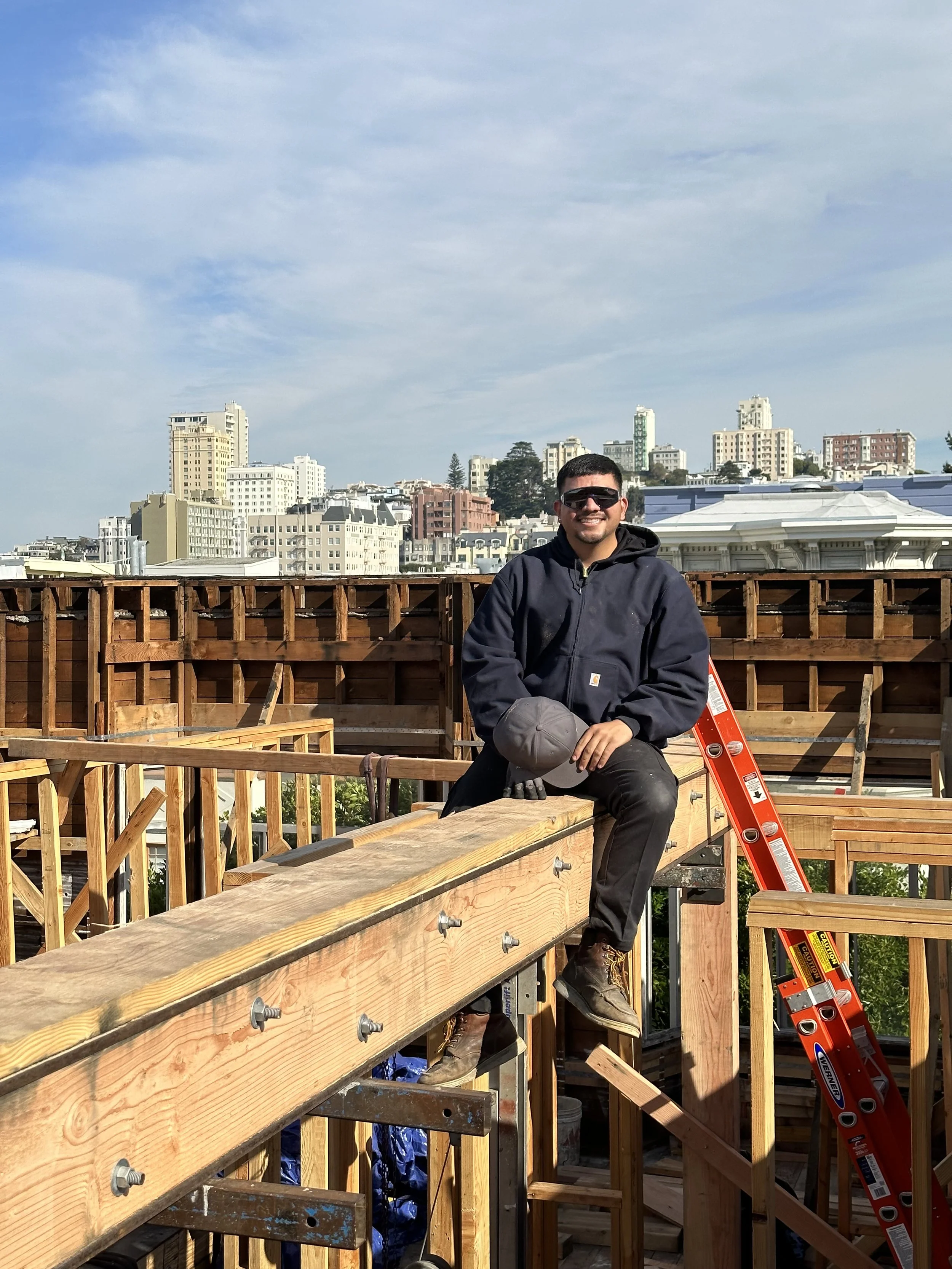A man wearing safety glasses and a dark jacket sitting on a construction site with wooden framing, a red ladder leaning against the structure, and a cityscape with tall buildings in the background.