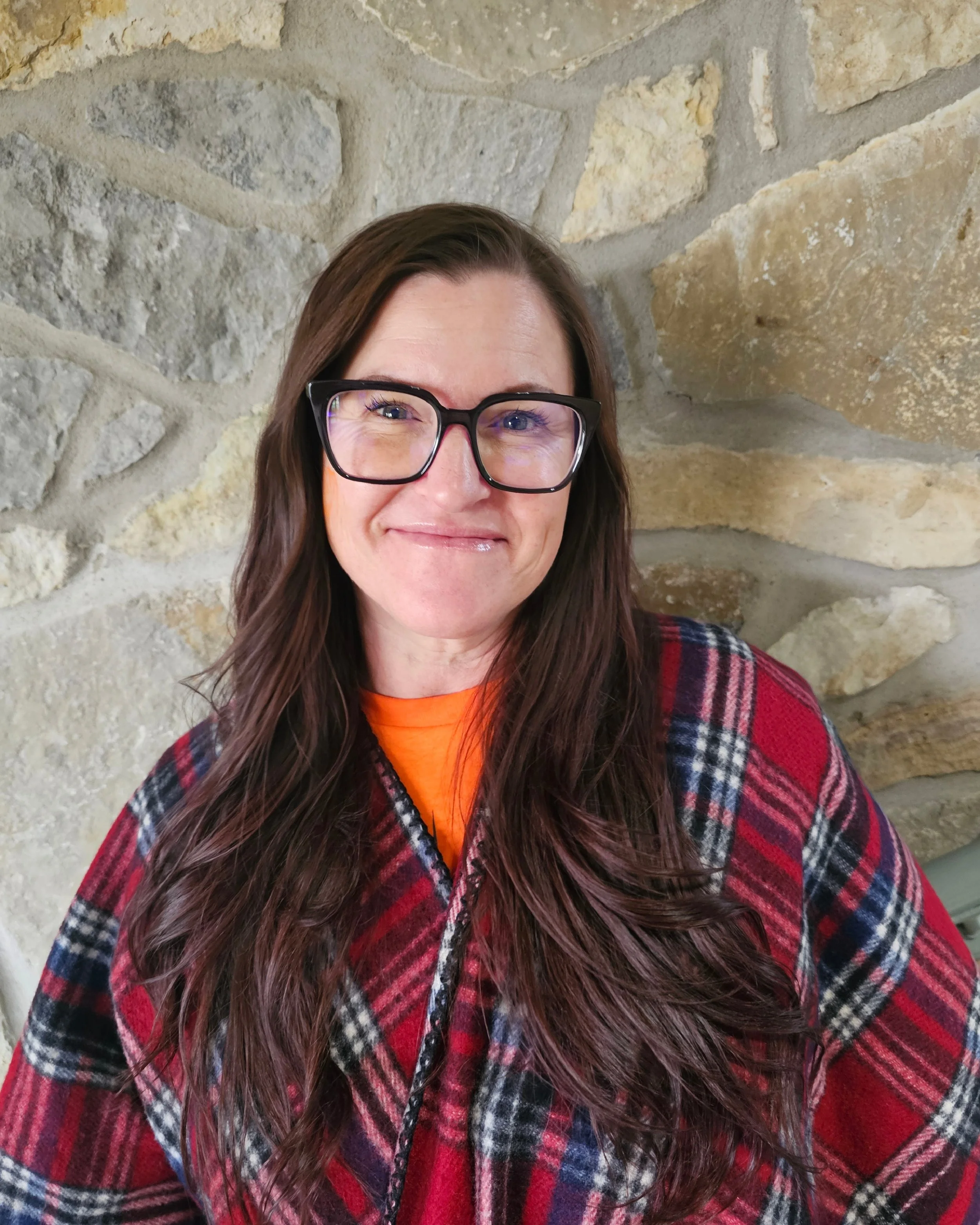 A woman with long brown hair, wearing glasses, a red plaid jacket, and an orange shirt, smiling in front of a stone wall.