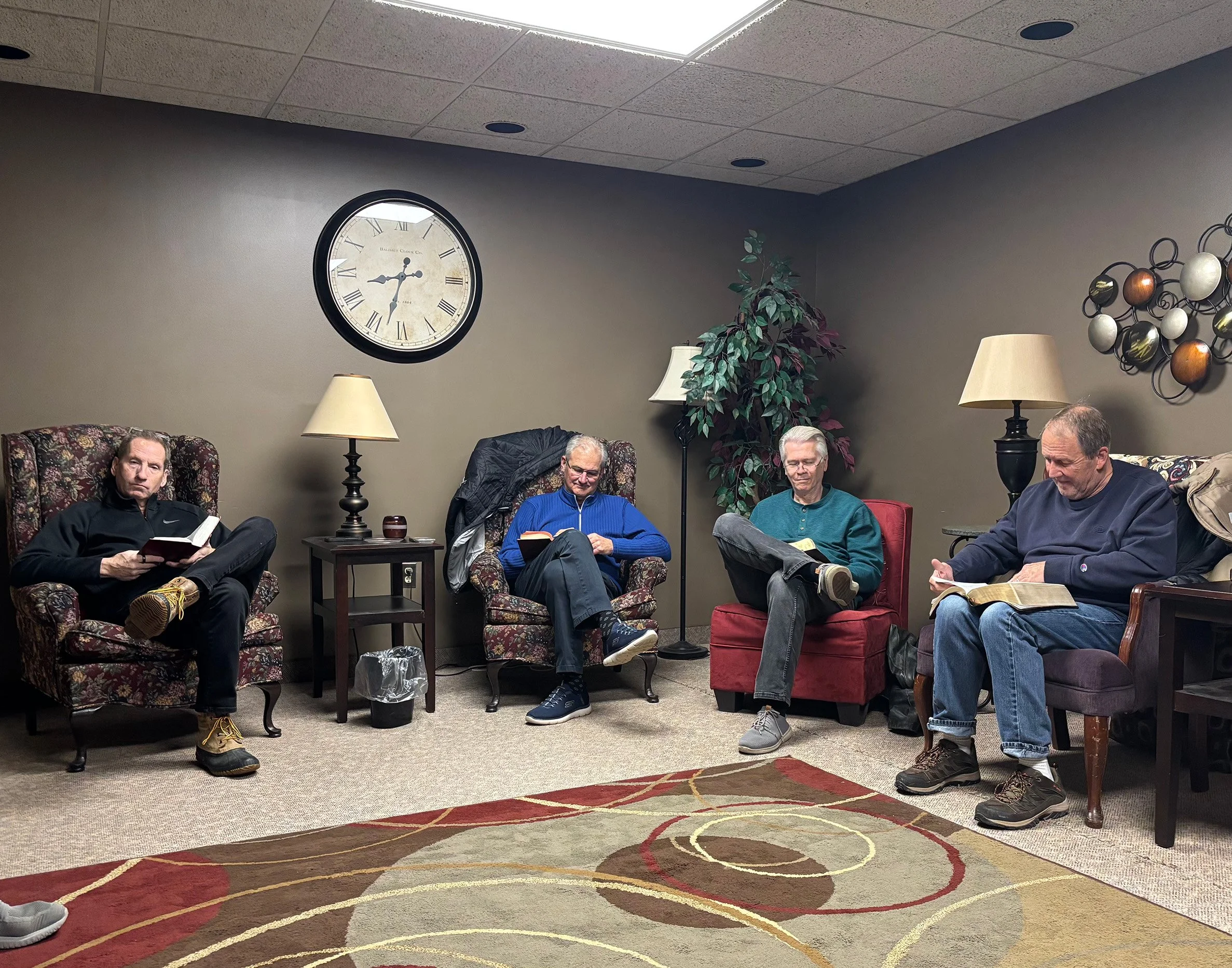 Four men sitting in a waiting room, reading books or magazines. The room has beige walls, a large clock showing 8:15, two lamps, plants, and wall art.